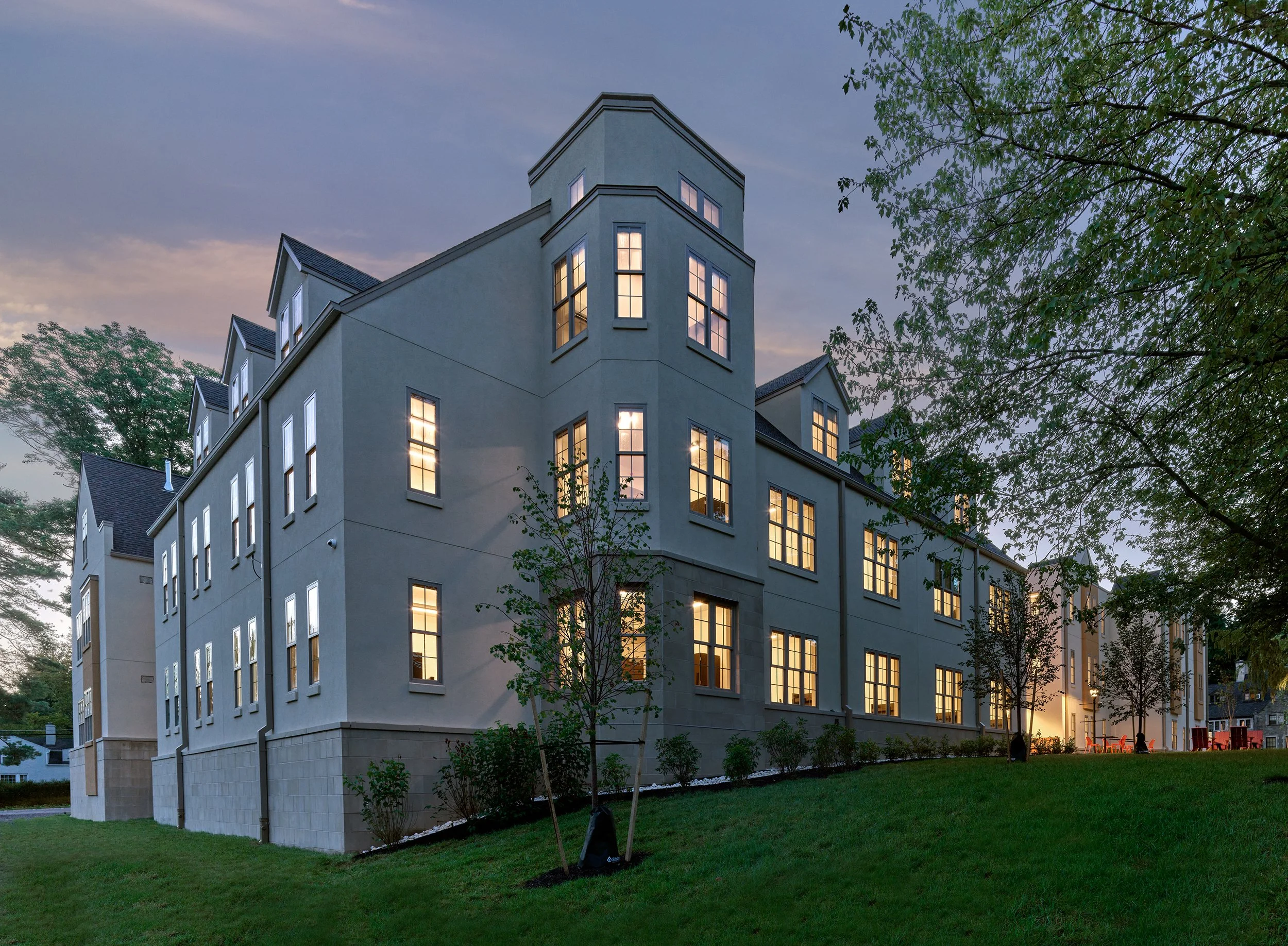 A multi-story residence hall with large lit windows at dusk, surrounded by a grassy lawn and trees.