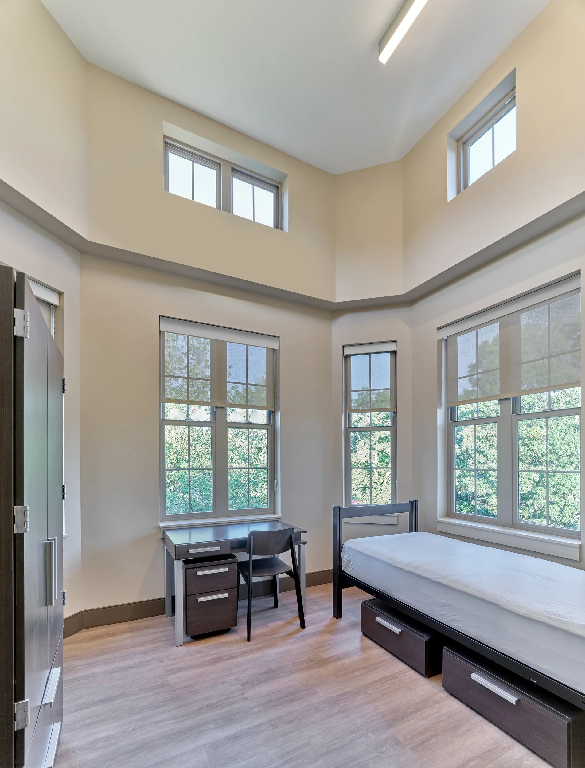 A bright dormitory room with high ceilings, multiple windows with blinds, a bed with drawers underneath, a small desk with a chair, and a locker, with wooden floors and beige walls.