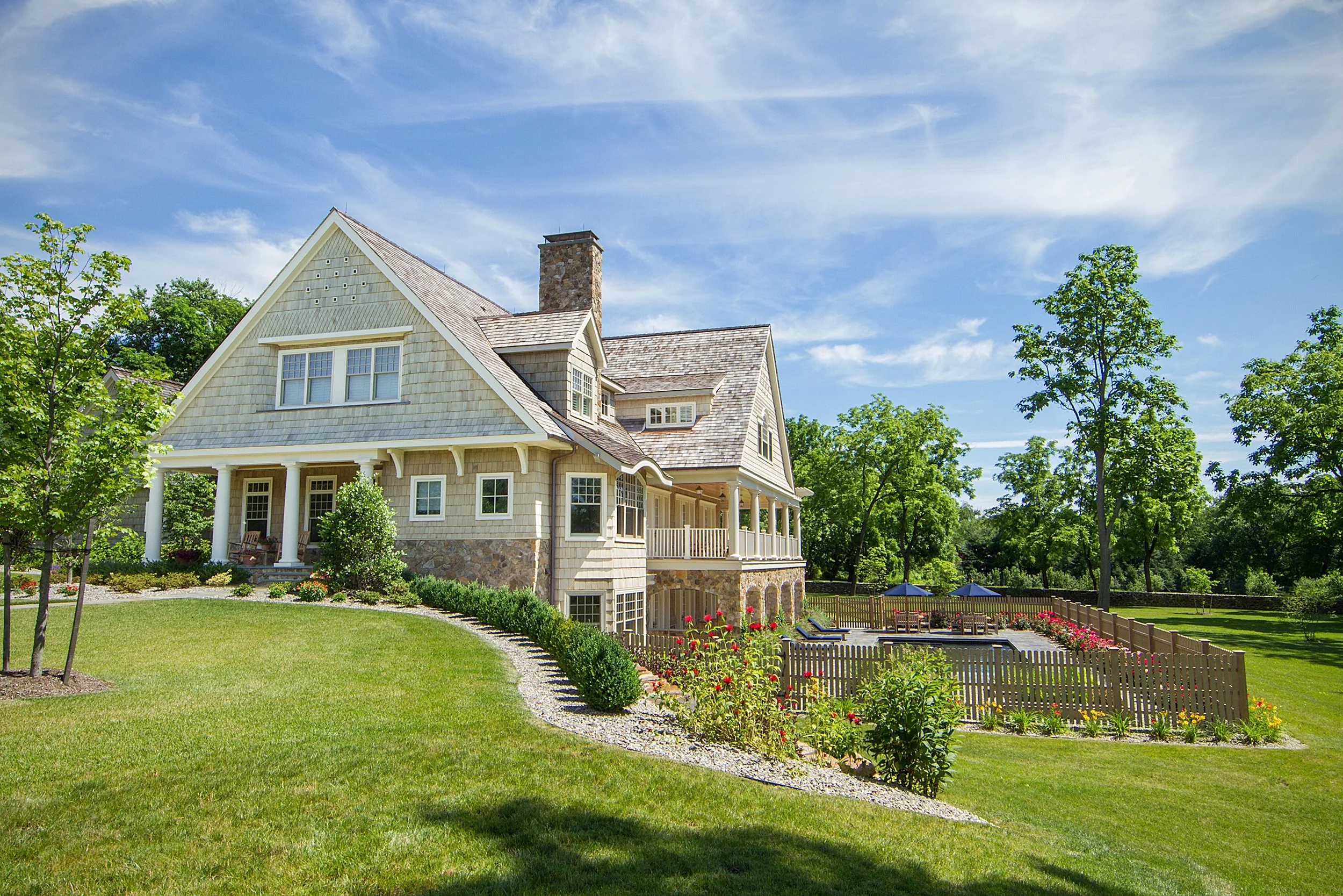 Front view of a large, beige house with a landscaped yard, green trees, and a wooden fence under a blue sky with some clouds.