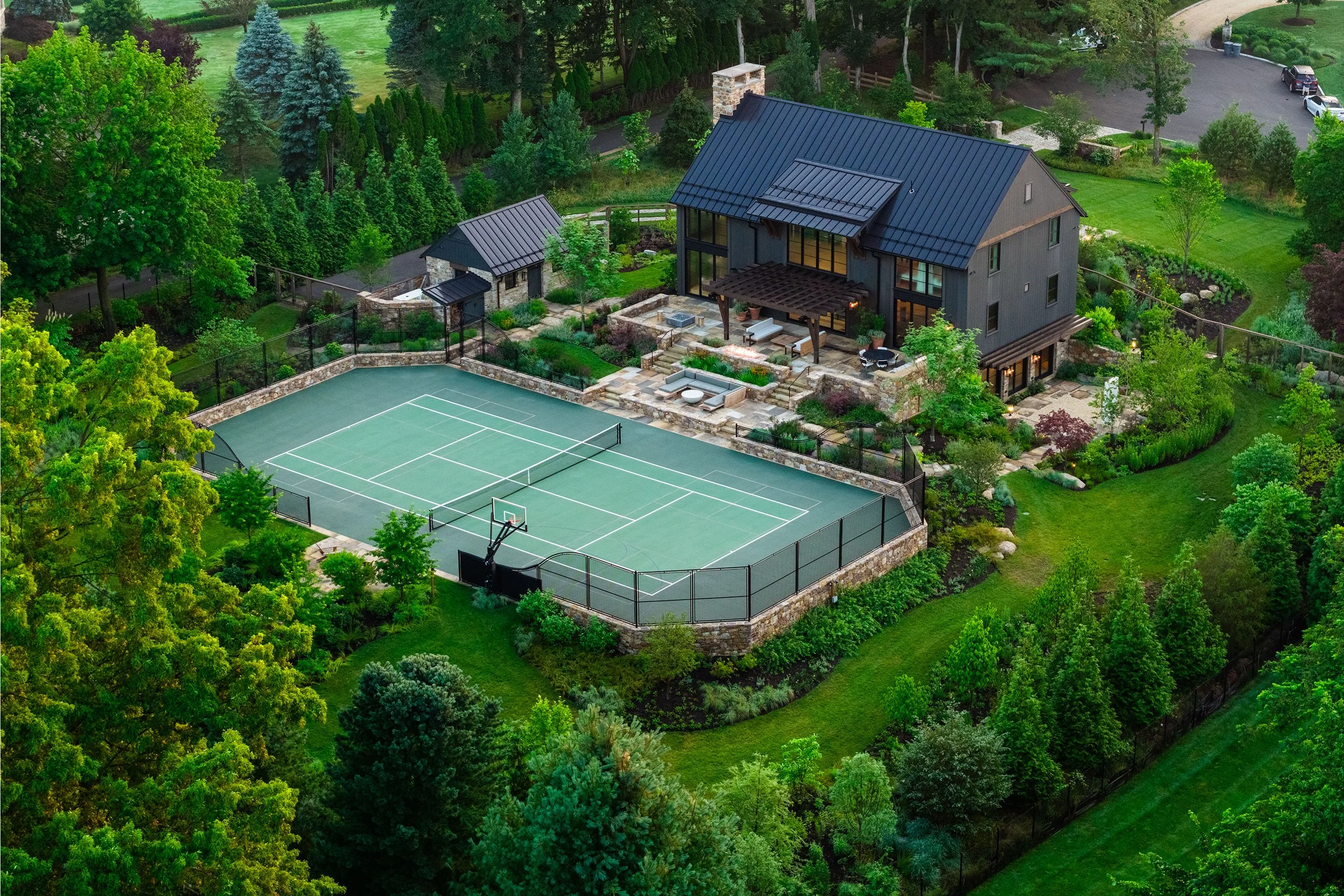 Aerial view of a tennis court, a modern barn with an outdoor lounge, gardens, and trees surrounding the area.