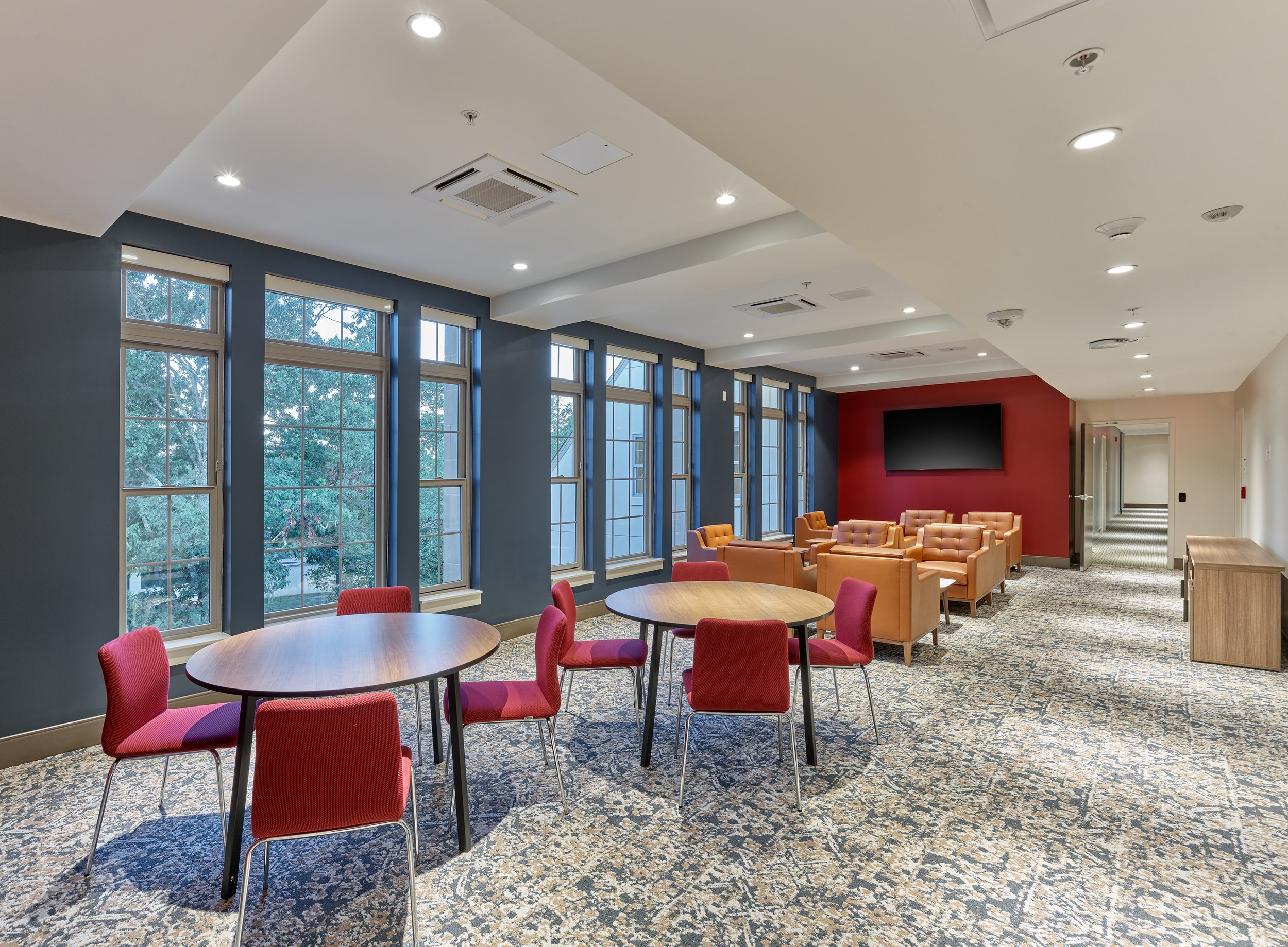 A spacious lounge area with large windows, round tables with red chairs, and a row of orange armchairs facing a wall-mounted TV. The room has a patterned carpet, blue accent walls, and a red wall behind the TV, with a hallway leading to another room.