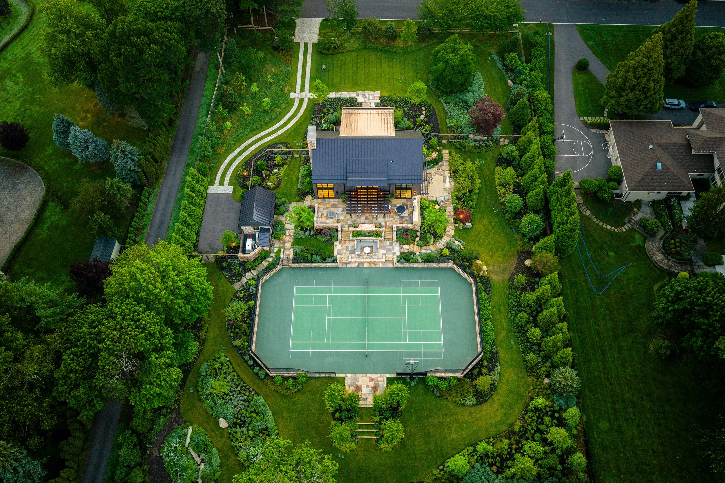 Aerial view of a tennis court, a modern barn, a stone pathway, and lush green landscaping and trees surrounding the area.