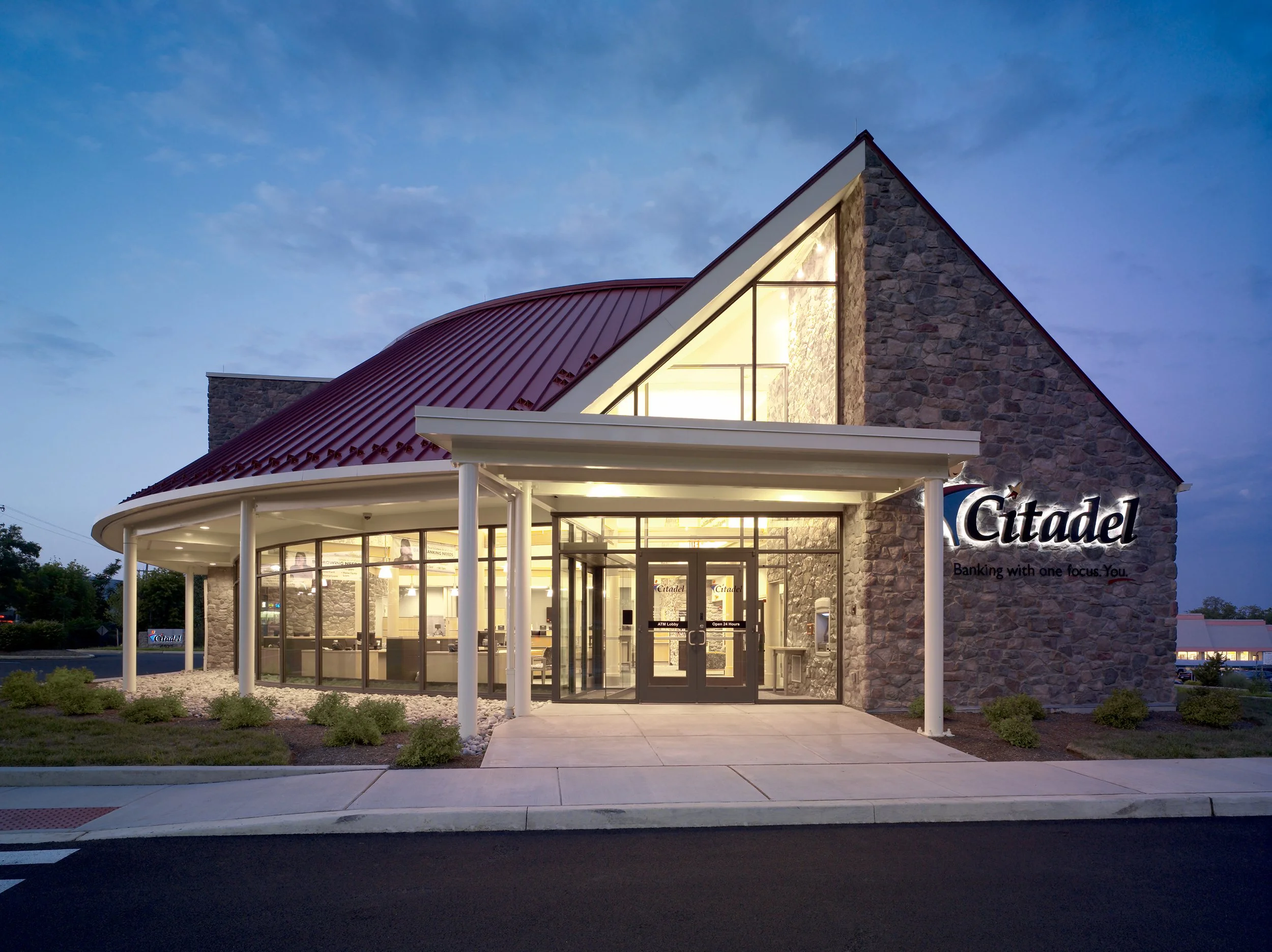 A modern bank building at dusk with a stone facade, large glass windows, and a maroon metal roof. The sign on the building reads 'Citadel' with the tagline 'Banking with one focus. You.' The entrance is illuminated, and there are small bushes and a s