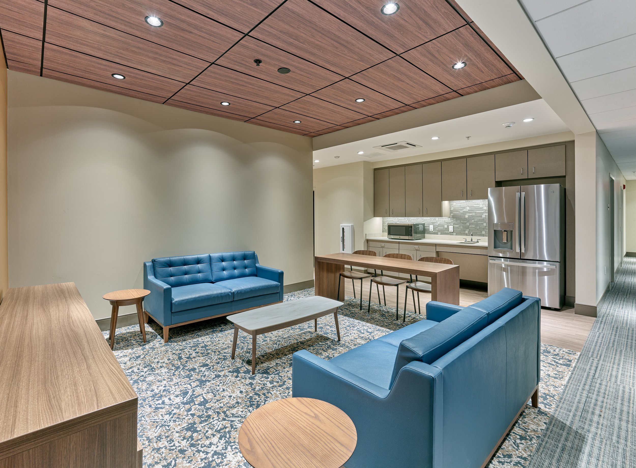 Modern dormitory kitchenette and lounge area with blue sofas, wooden tables, kitchen appliances, and a patterned rug.