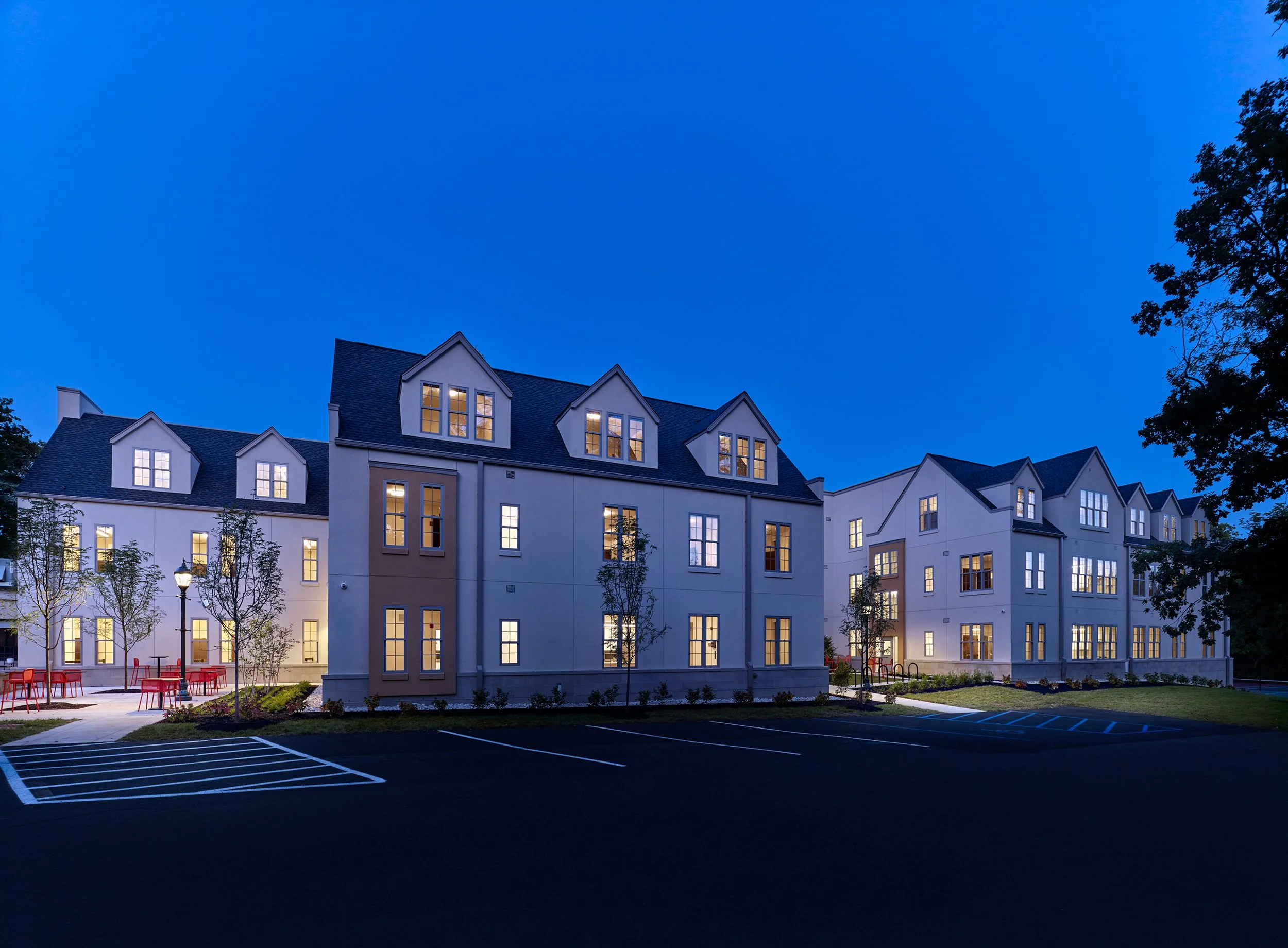 A modern multi-story residence hall at dusk with illuminated windows, surrounded by a parking lot, trees, and outdoor seating.