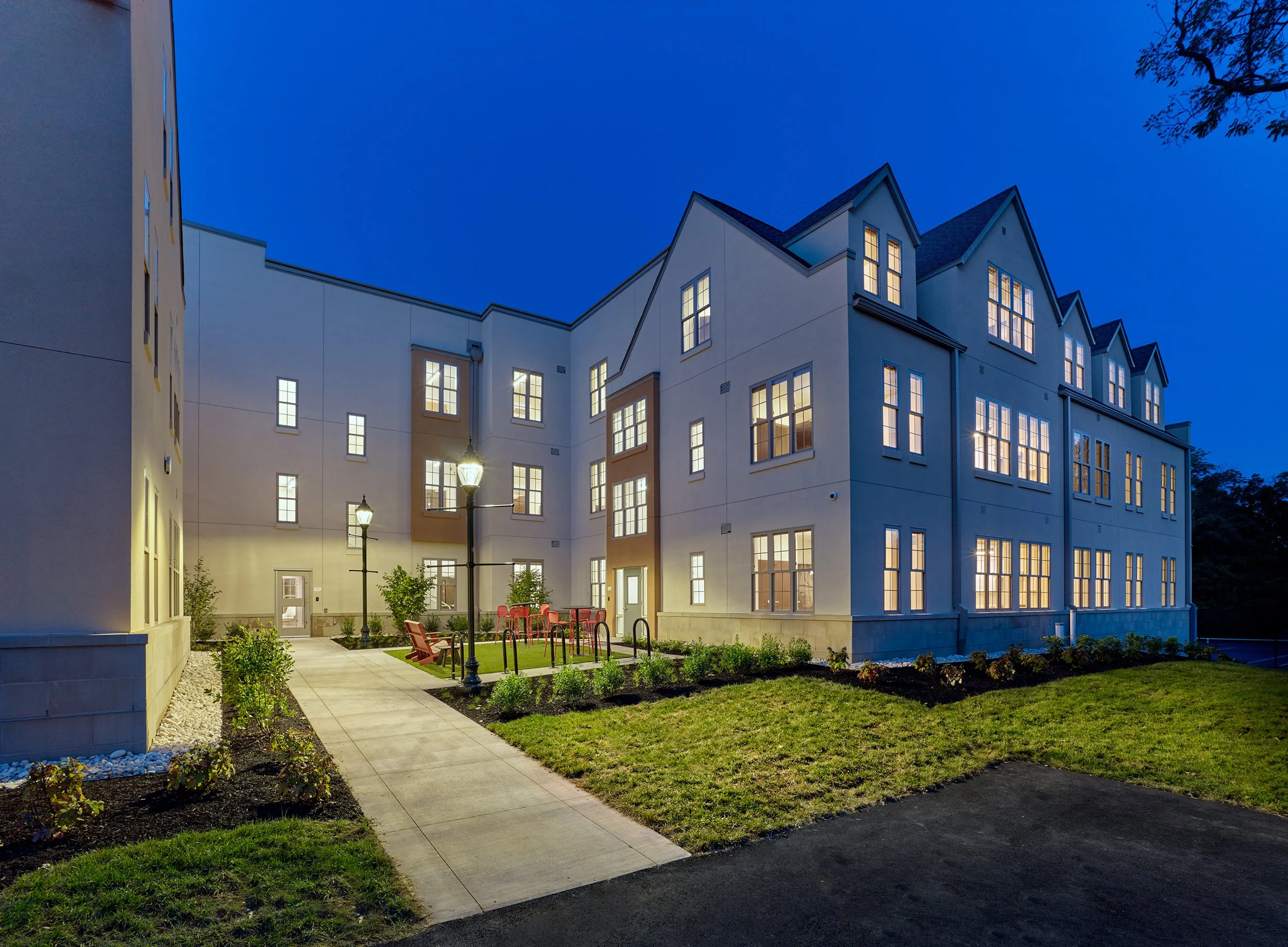 An residence hall at night with lit windows, a pathway, benches, and landscaped yard.