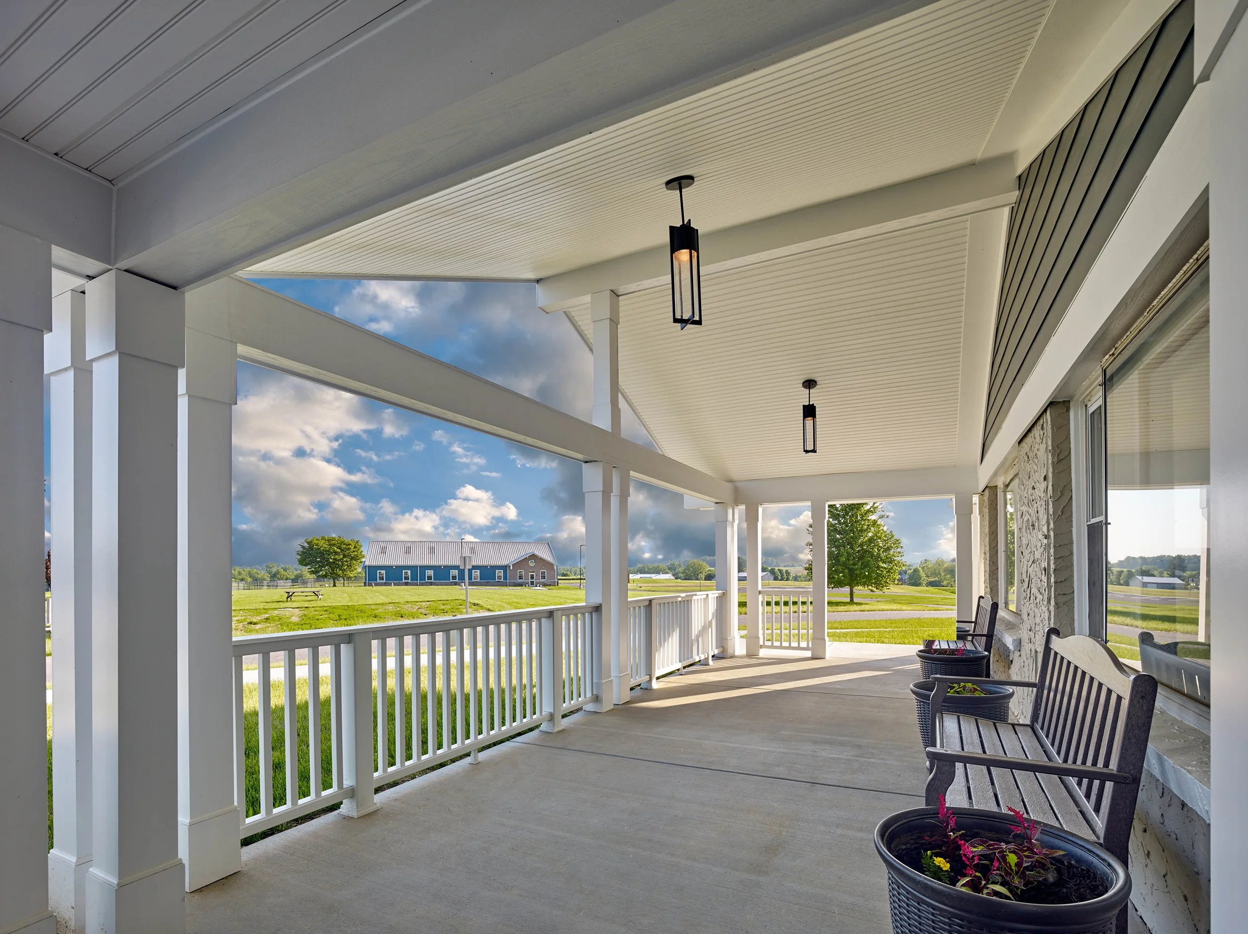 Covered porch with white railing, hanging light fixtures, and patio chairs overlooking a green grassy yard with a blue building in the distance under a partly cloudy sky.