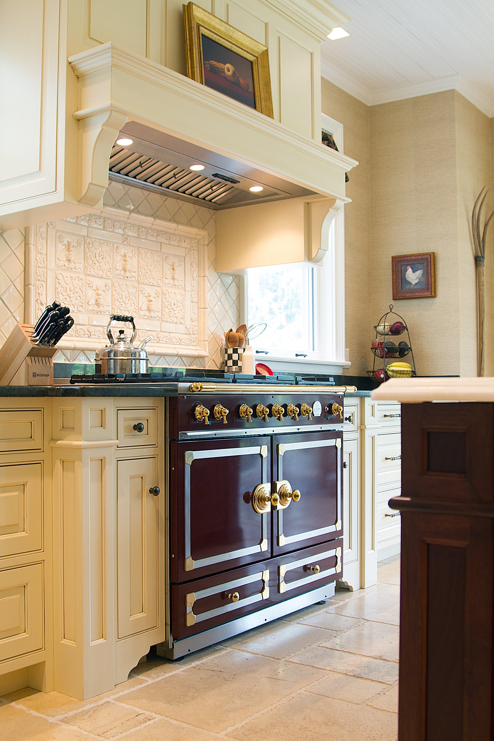 A kitchen with beige cabinetry, a black and gold stove, decorative wall tiles, a window, and framed pictures on the wall.