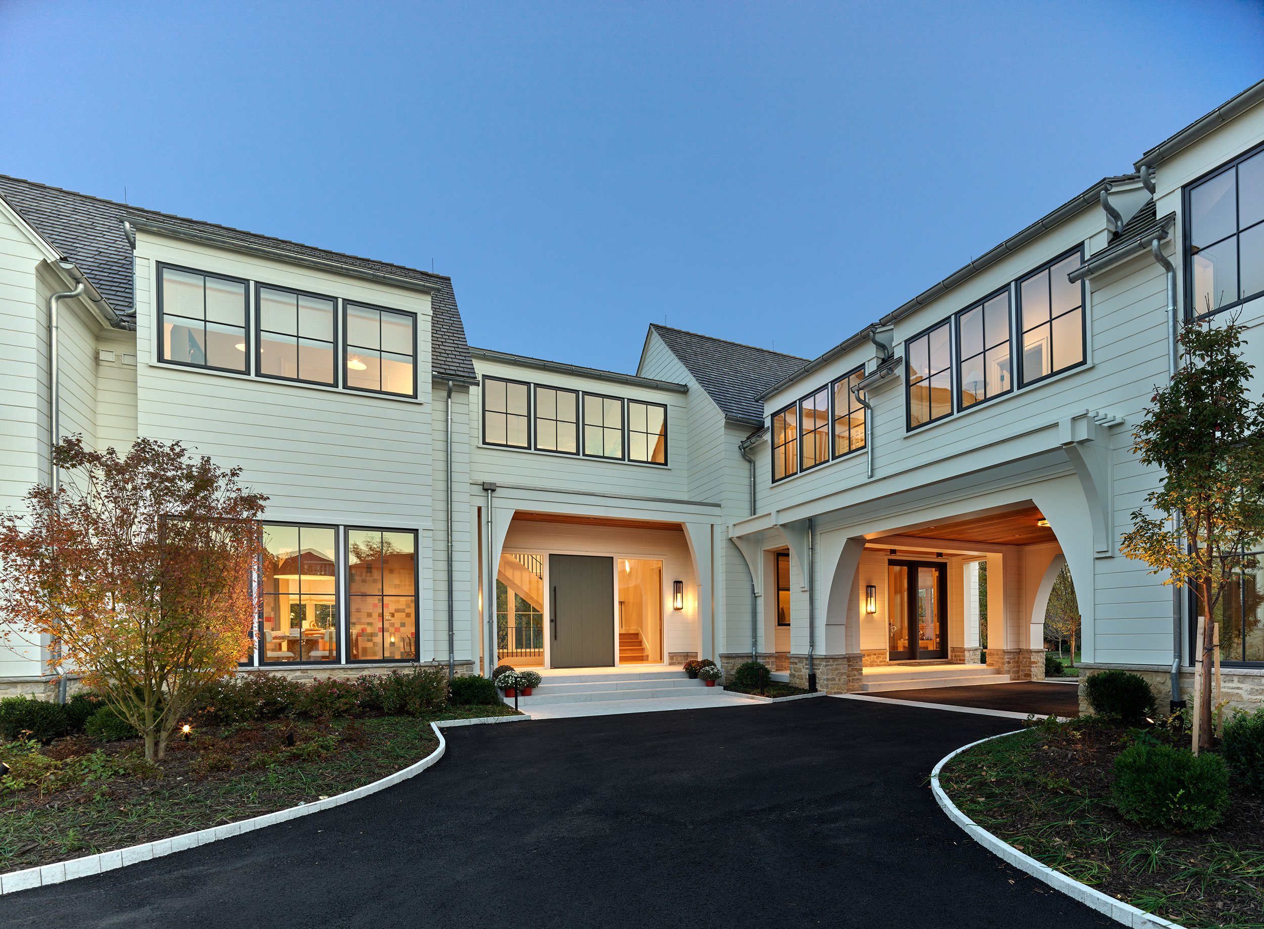 Modern multi-story white house with large windows, stone foundation, large front door with porte-cochere to the right, and steps leading to the entrance decorated with potted chrysanthemums, at dusk.