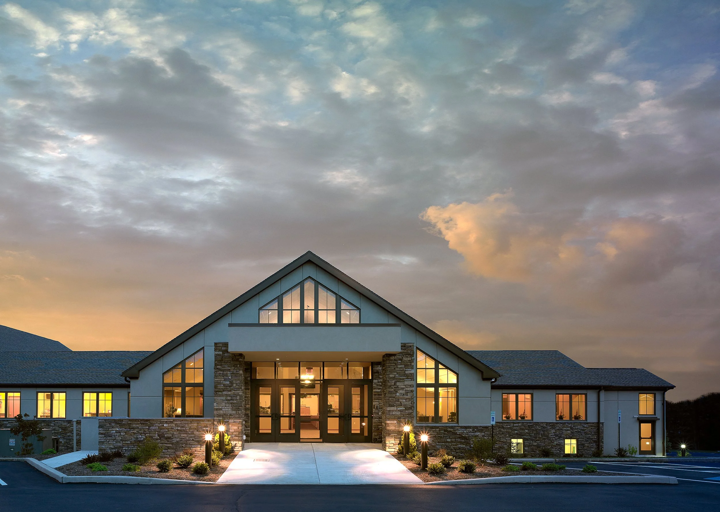 Modern church building with stone accents and large windows, illuminated at sunset, with a parking lot and landscaped bushes in front.