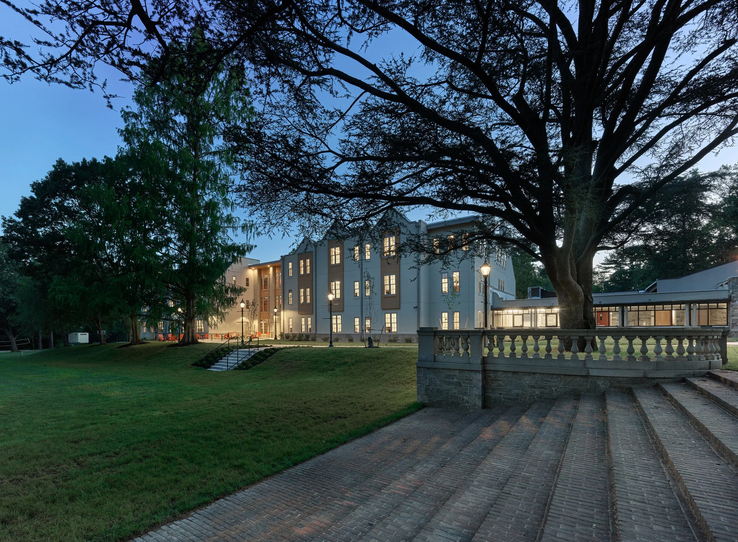 A modern multi-story dormitory with illuminated windows surrounded by trees and green lawn, taken during evening or twilight.