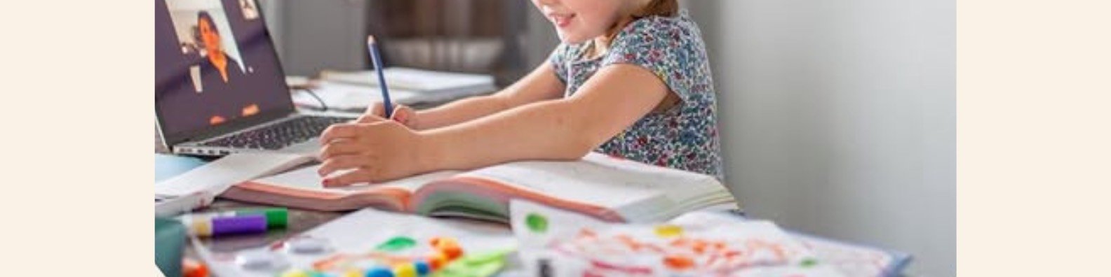 A young girl sitting at a cluttered desk working on a craft or school project, with an open book, colorful markers, and craft supplies, while participating in a virtual meeting or class on her laptop.