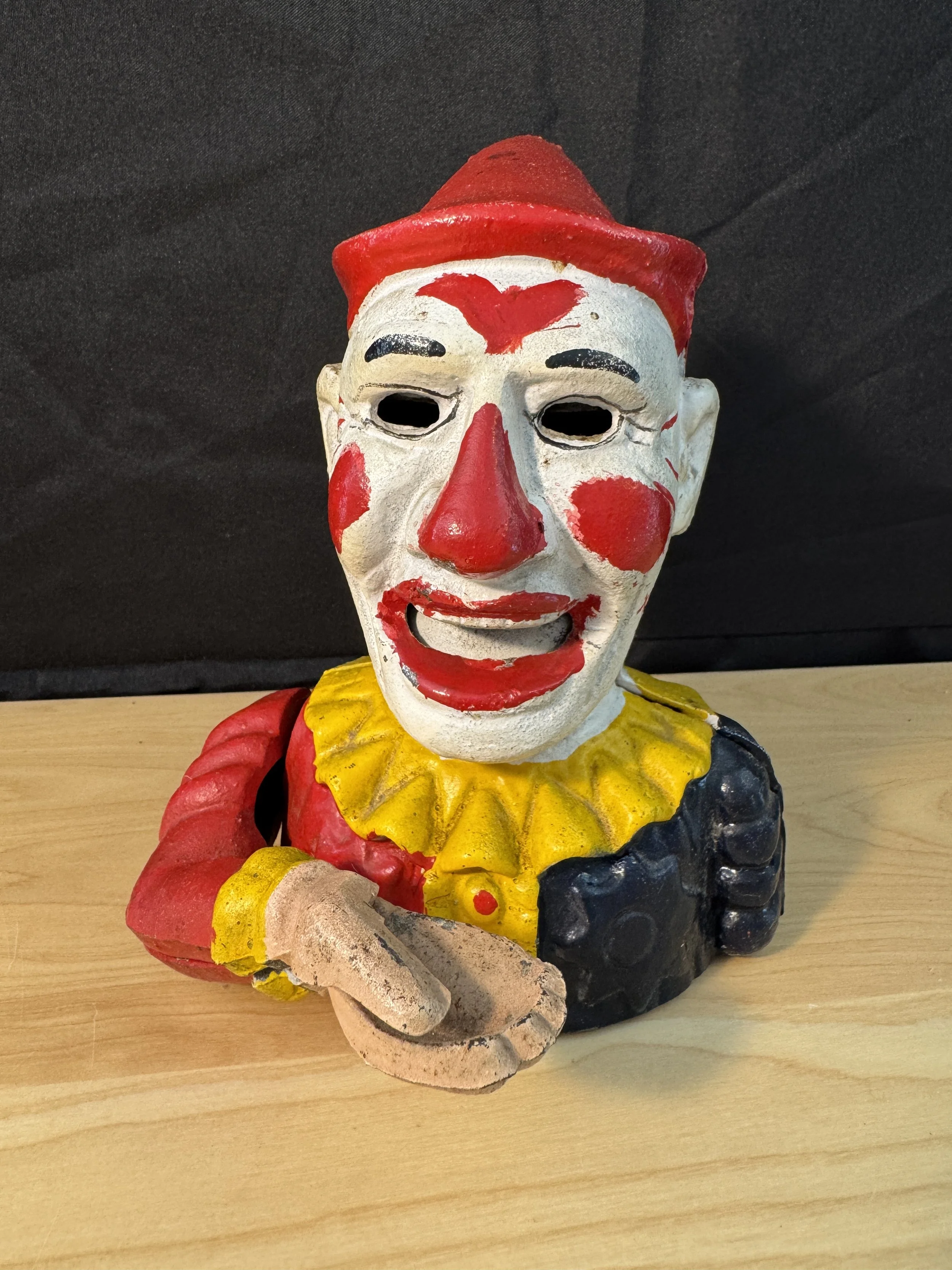 Colorful clown figurine with red hair, white face, red and black facial features, yellow collar, and red, black, and beige clothing, standing on a wooden surface against a black background.