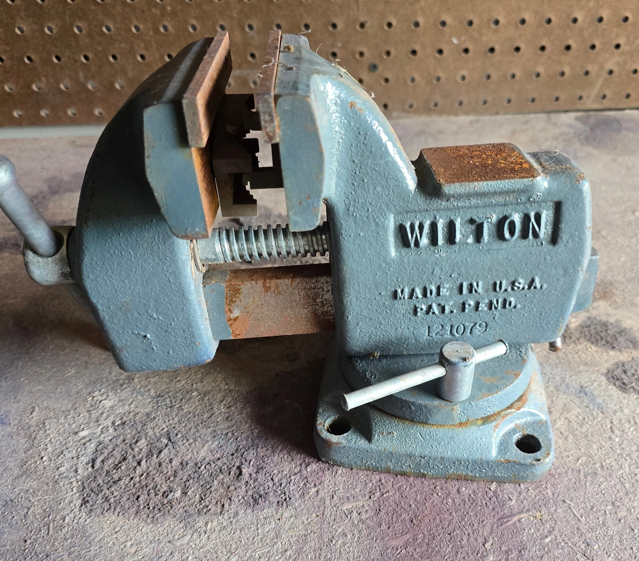 A large blue Wilton bench vise with rust on its handle and contact points, mounted on a workbench with a pegboard in the background.