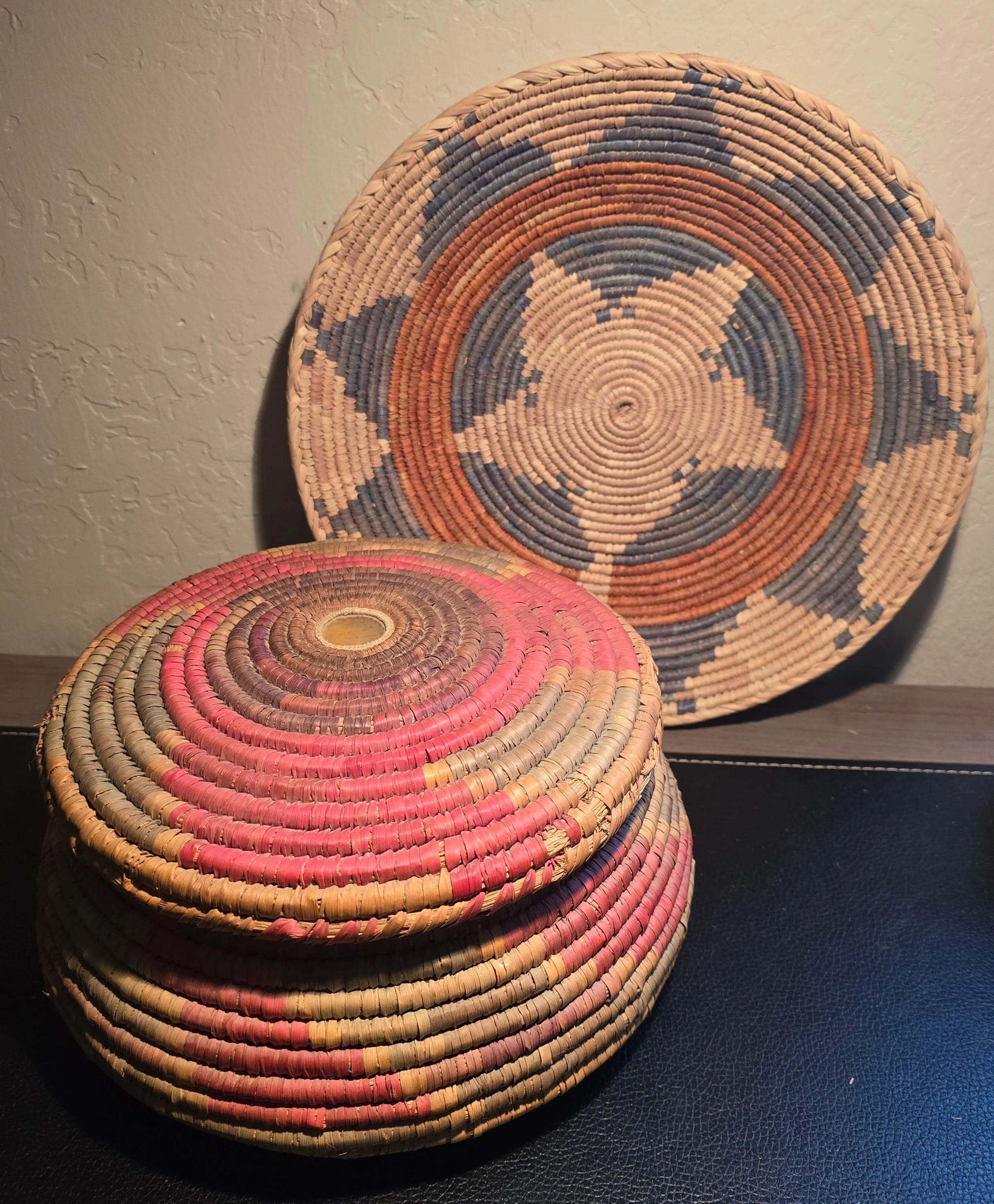 A collection of three woven baskets, with the largest in the background, medium in the middle, and smallest in the foreground. The baskets feature a spiral pattern with colors including beige, red, orange, blue, and pink. The background is a textured beige wall and the surface is dark.