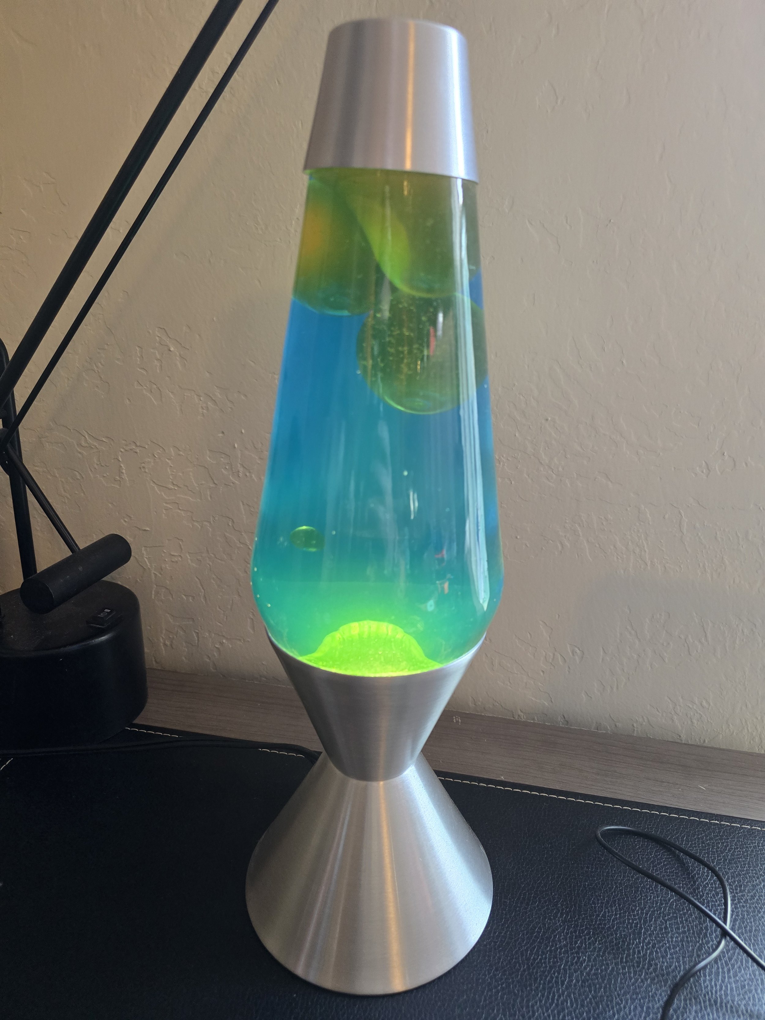 A lava lamp with blue liquid and green wax inside, placed on a black surface near a textured wall, with part of a desk lamp visible on the left and a power cord on the right.