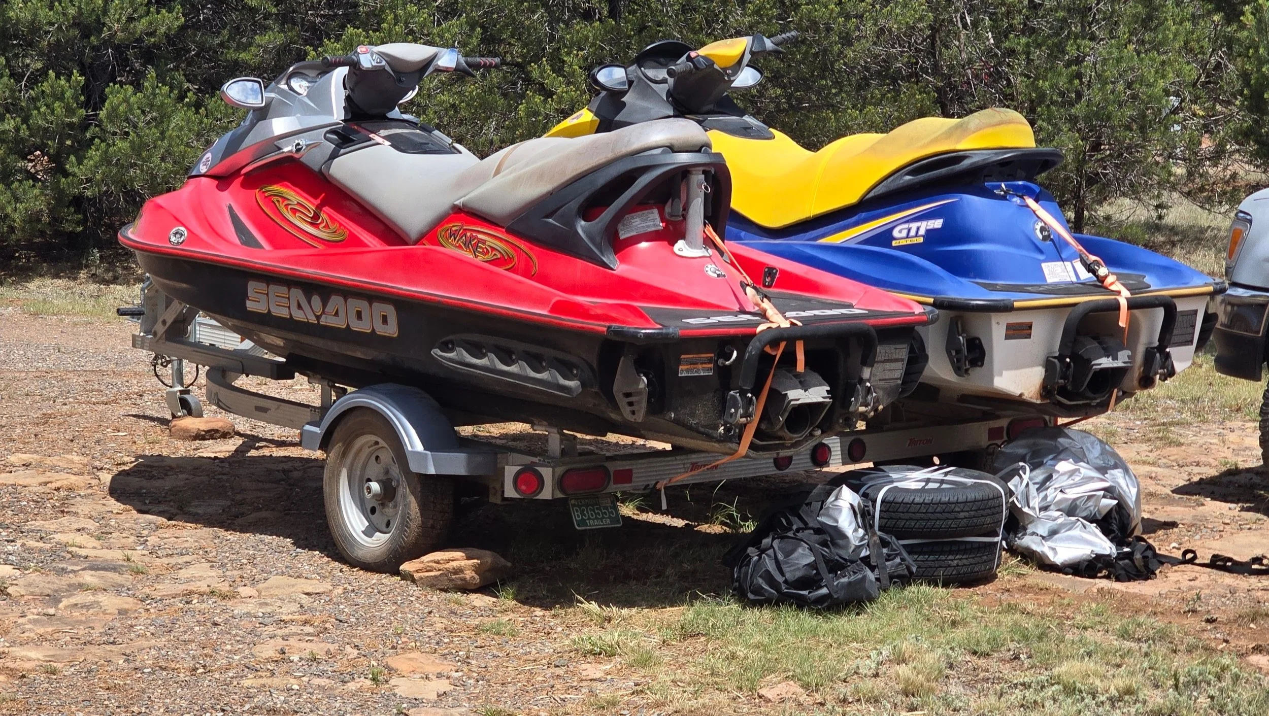 A trailer with two jet skis parked on rocky ground, surrounded by bushes. One jet ski is red, and the other is blue and yellow. There are bags and a spare tire on the ground nearby.
