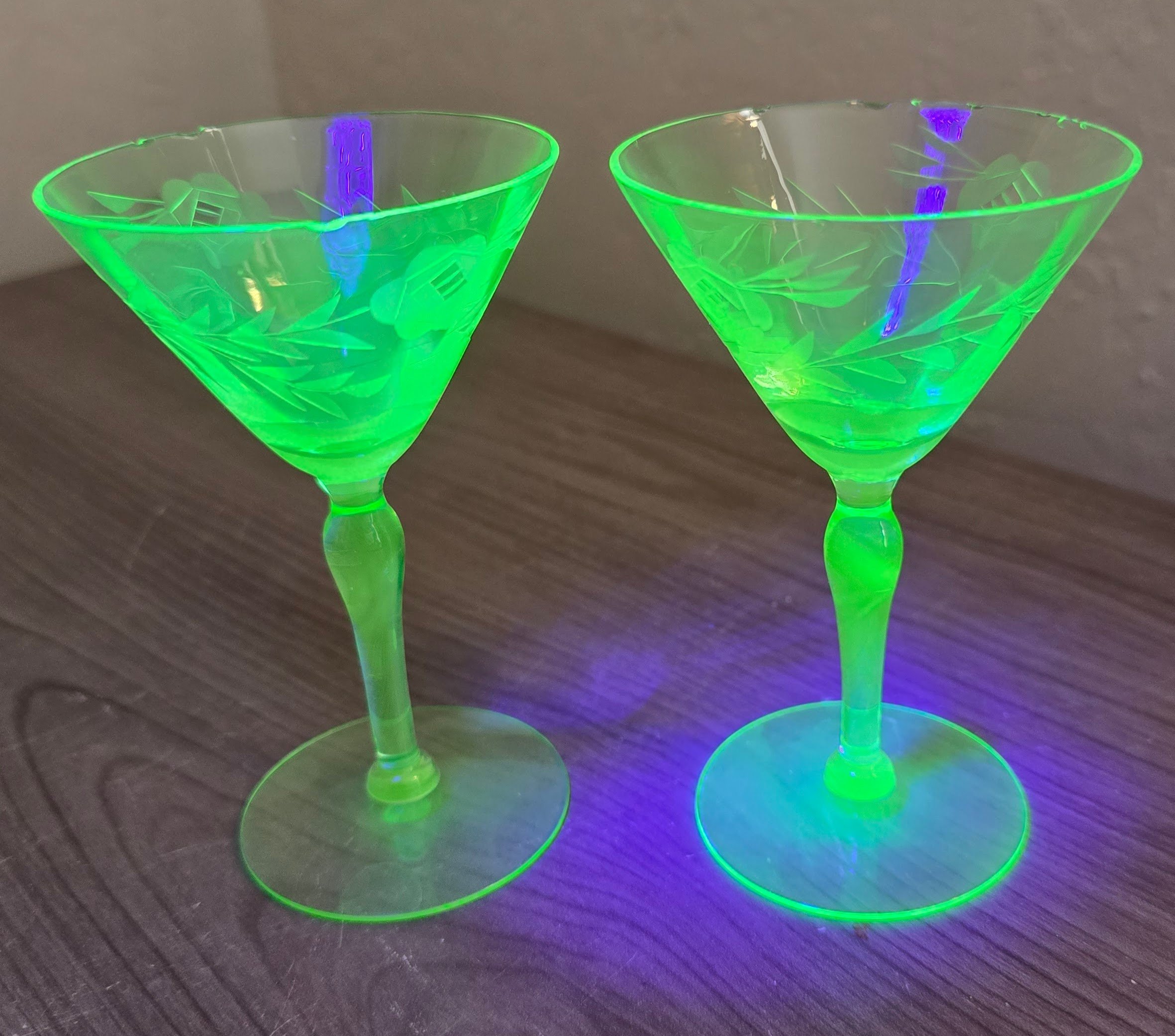 Two transparent cocktail glasses with green and purple glow-in-the-dark designs on a dark surface.