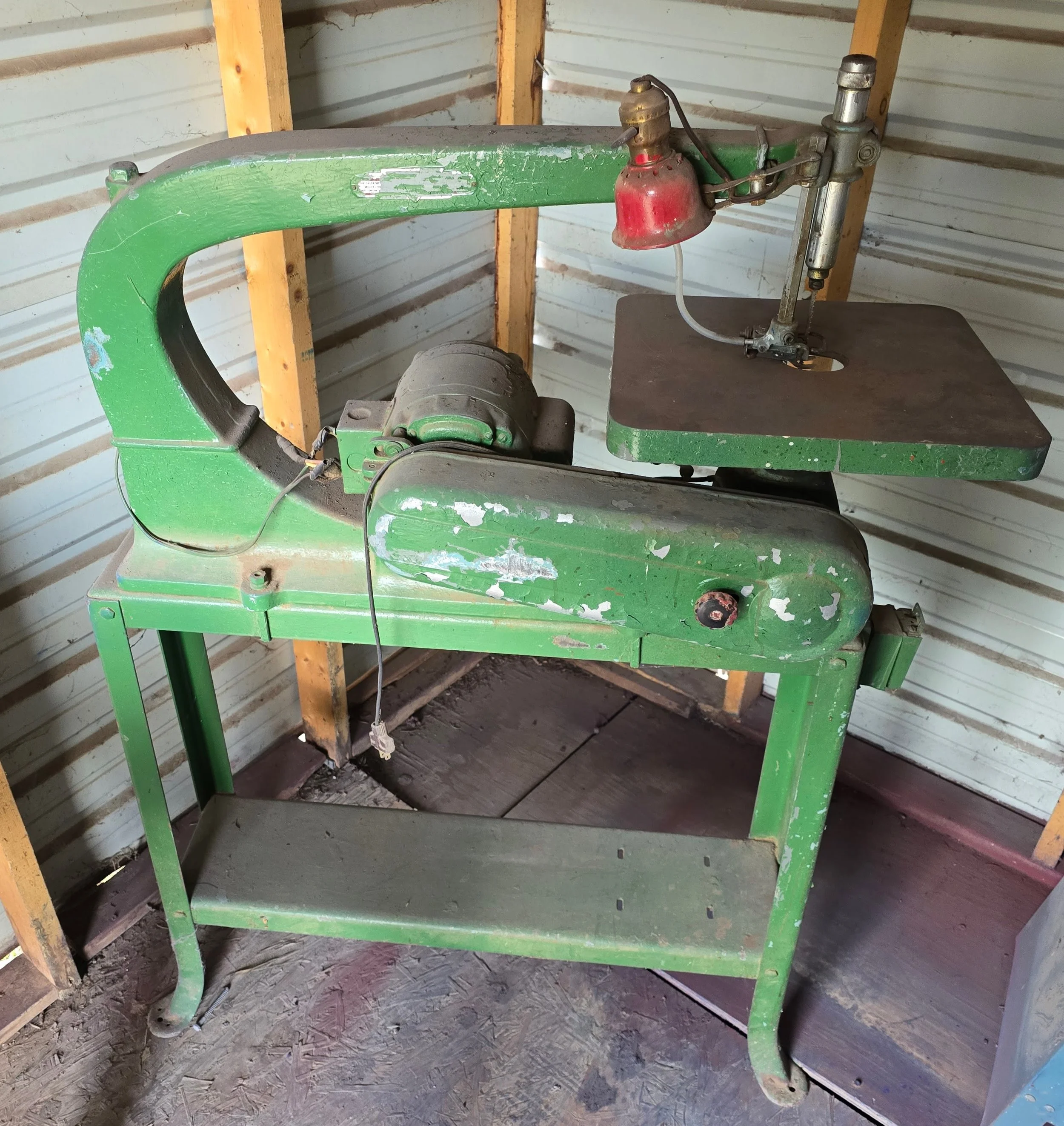 An old green bandsaw with a red safety guard and a silver blade, situated in a wooden shed with metal siding. The paint on the bandsaw is chipped and worn.