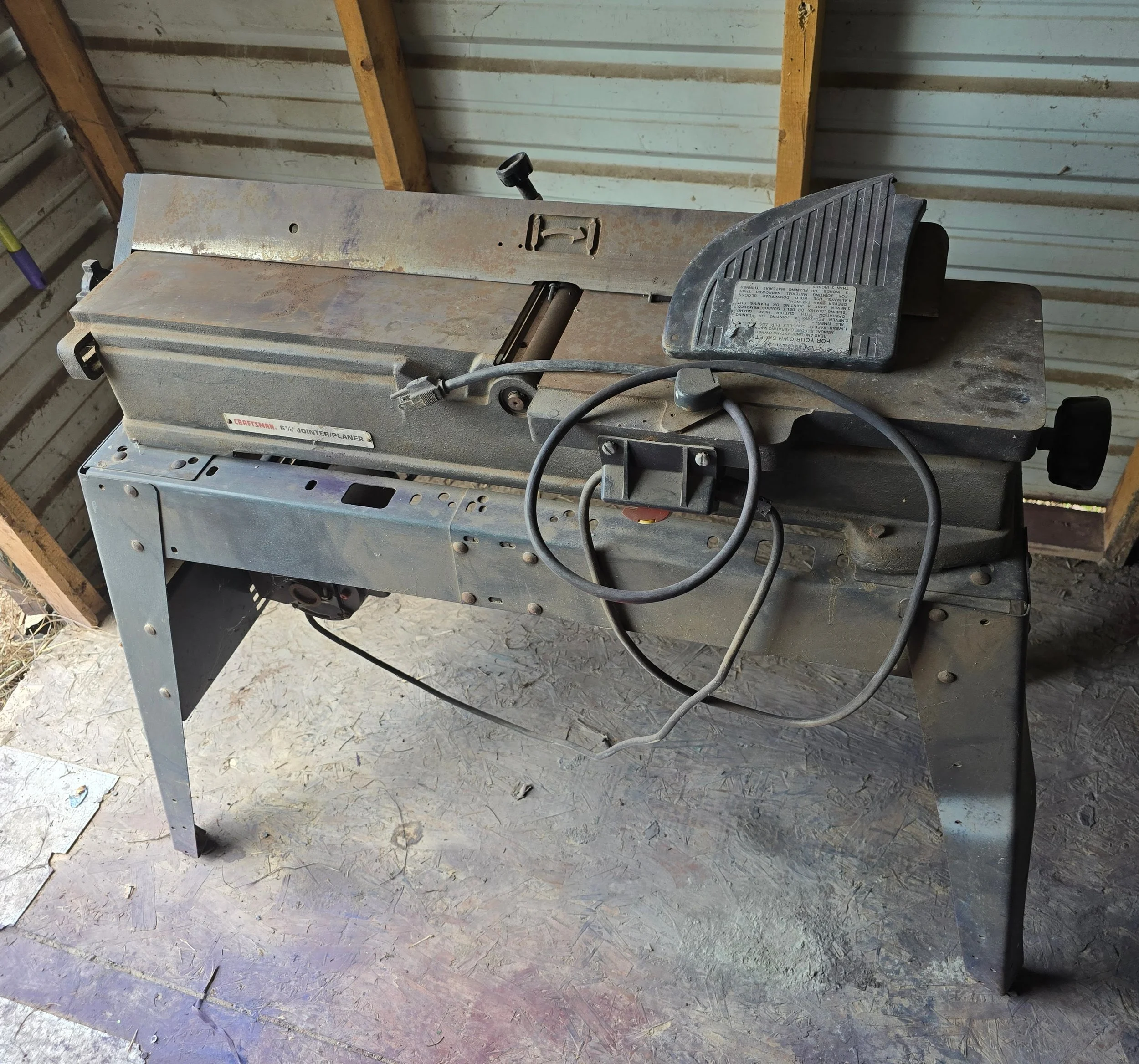 A metal table saw with a black push stick resting on it, placed on a wooden floor in a workshop with metal walls and wooden support beams.