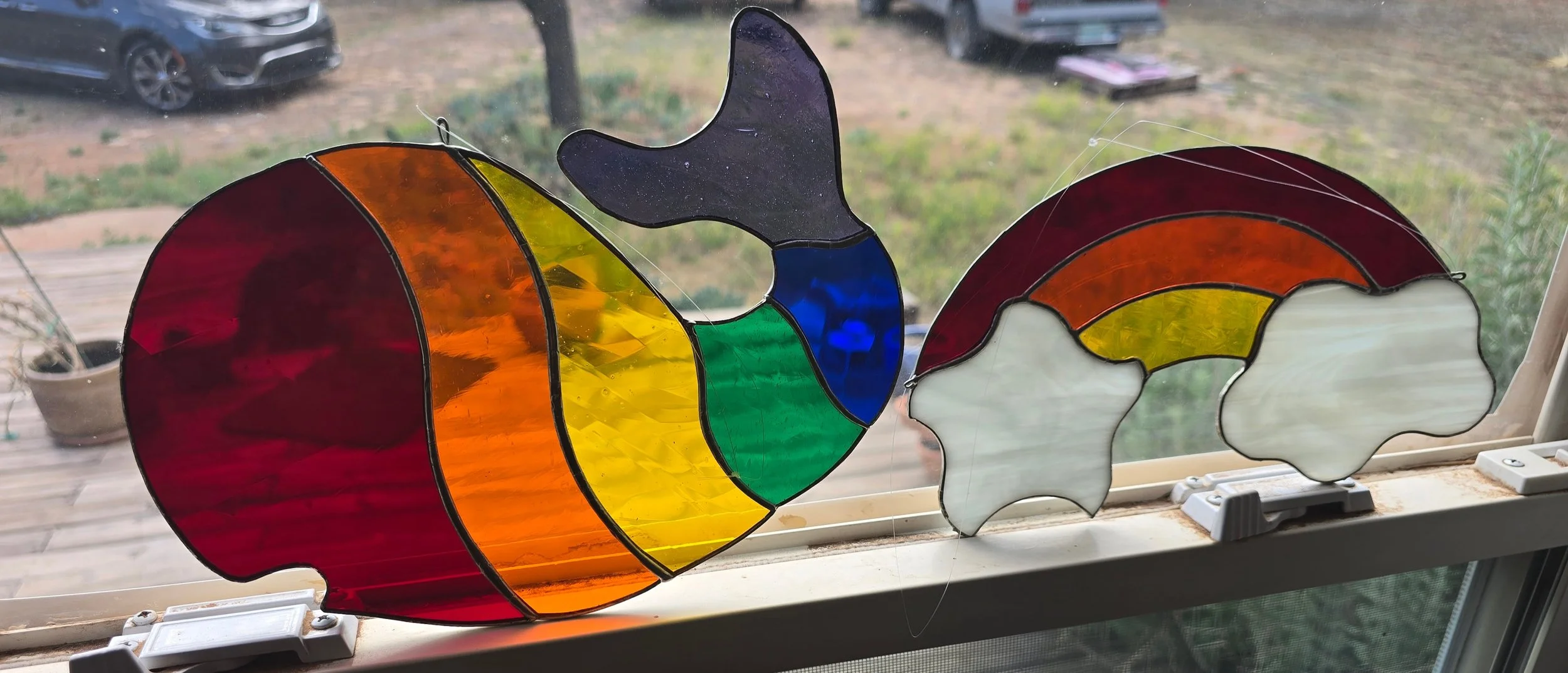 Colorful stained glass artwork depicting a rainbow with a black silhouette of a butterfly and white cloud shapes, displayed on a window with outdoor scenery in the background.