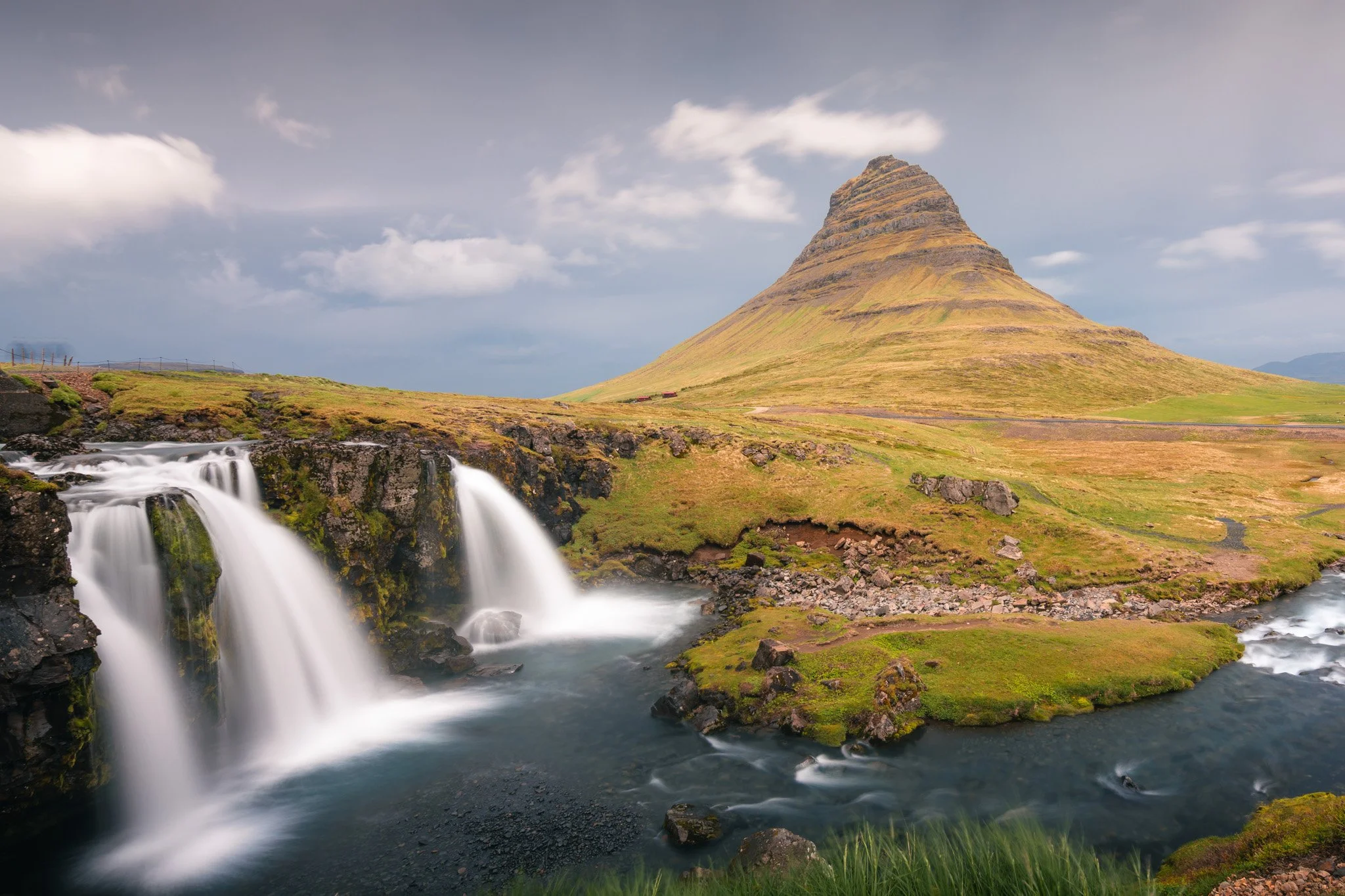 Scenic landscape of Kirkjufell mountain with waterfalls and river in the foreground, cloudy sky in the background.
