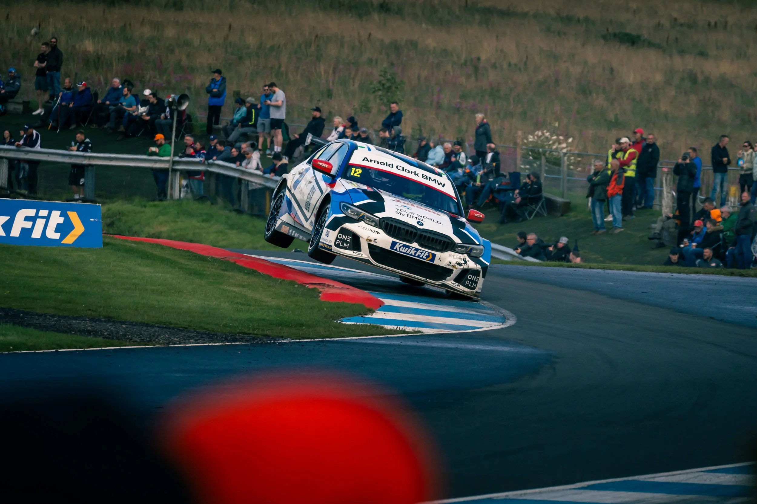 A race car is airborne while taking a sharp turn on a racetrack, with spectators watching from the sidelines.