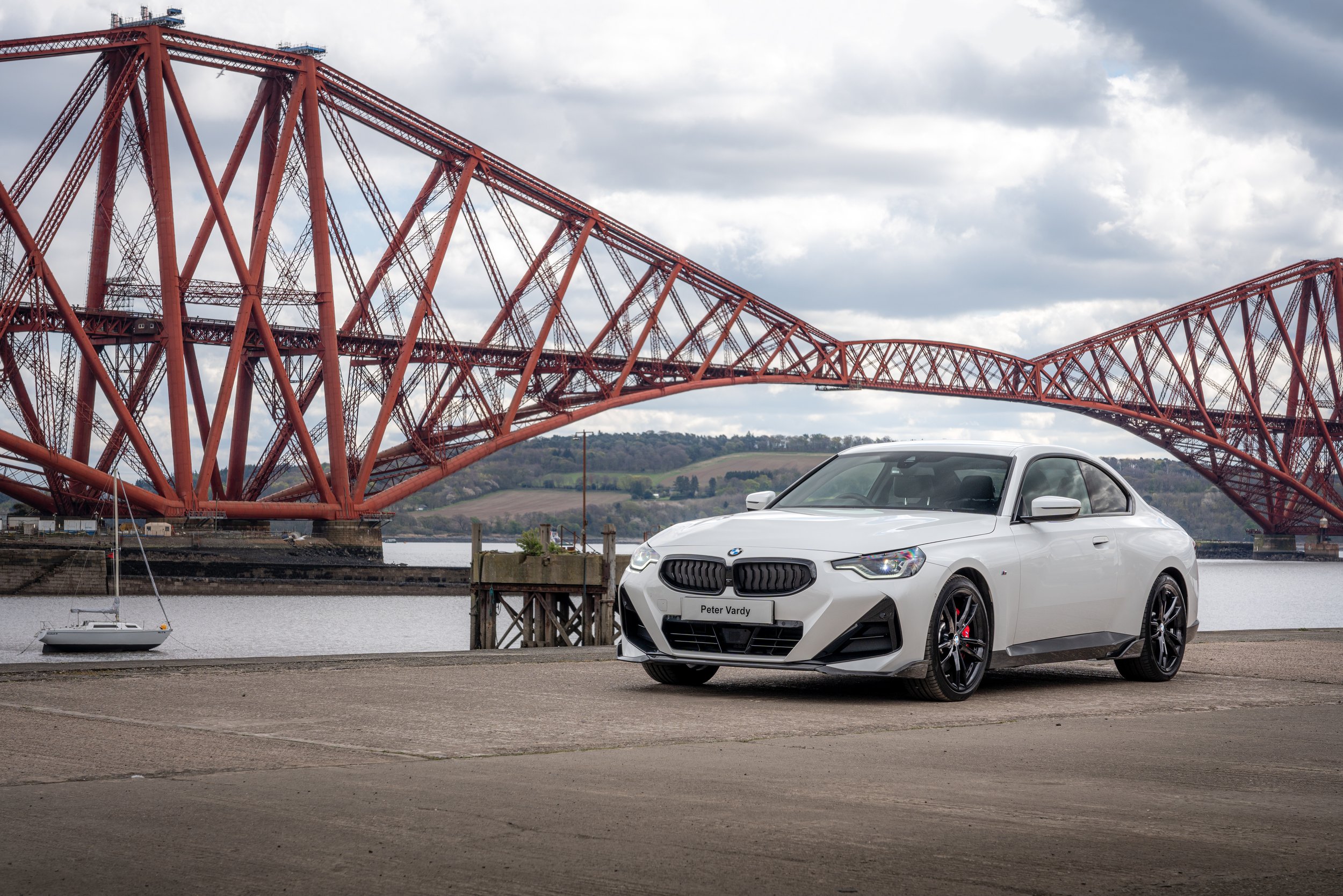 A white BMW car parked on a concrete surface near a body of water, with a large red steel bridge and cloudy sky in the background.