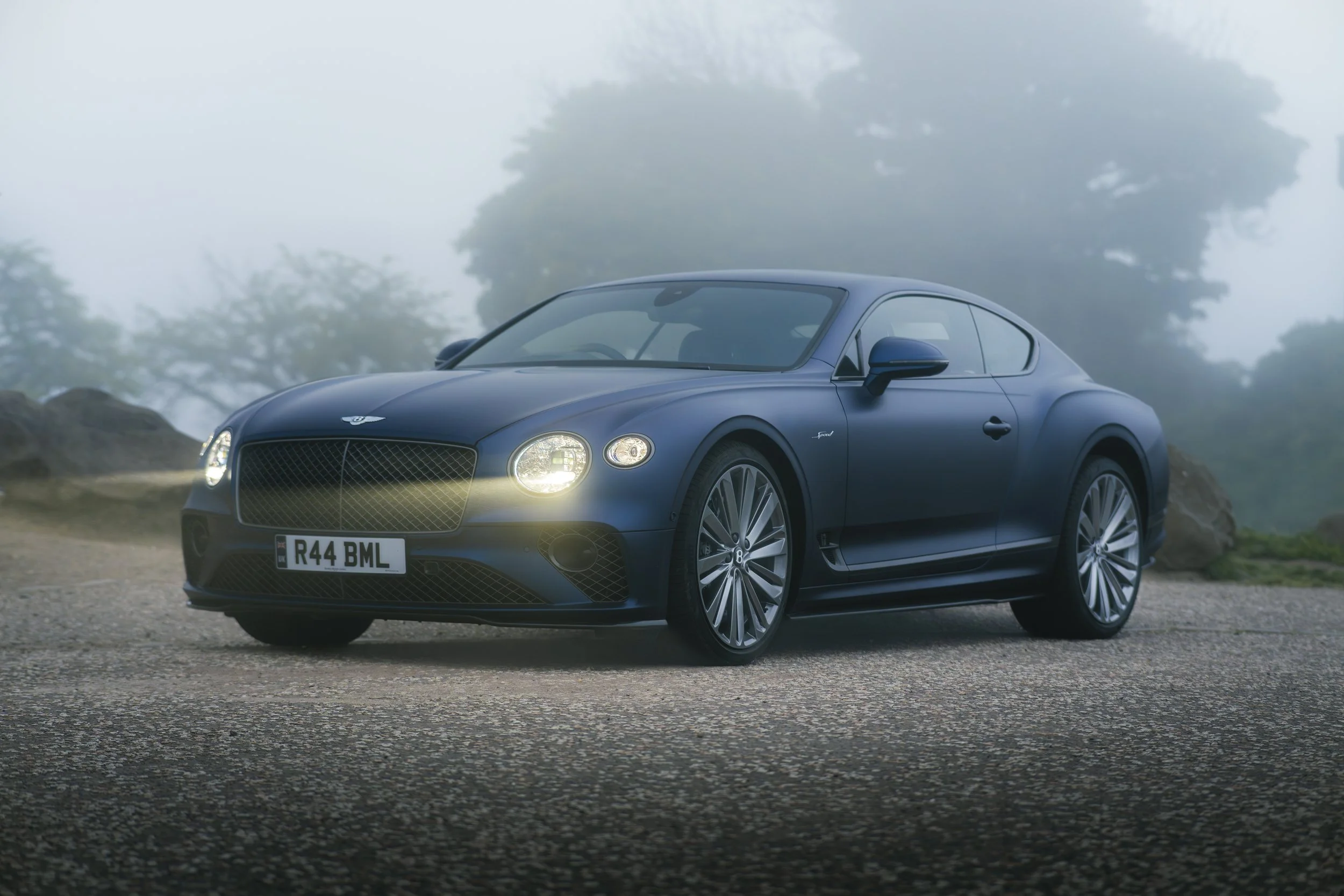 A dark blue Bentley Continental GT parked on a gravel surface, with foggy trees in the background and the car's headlight on.