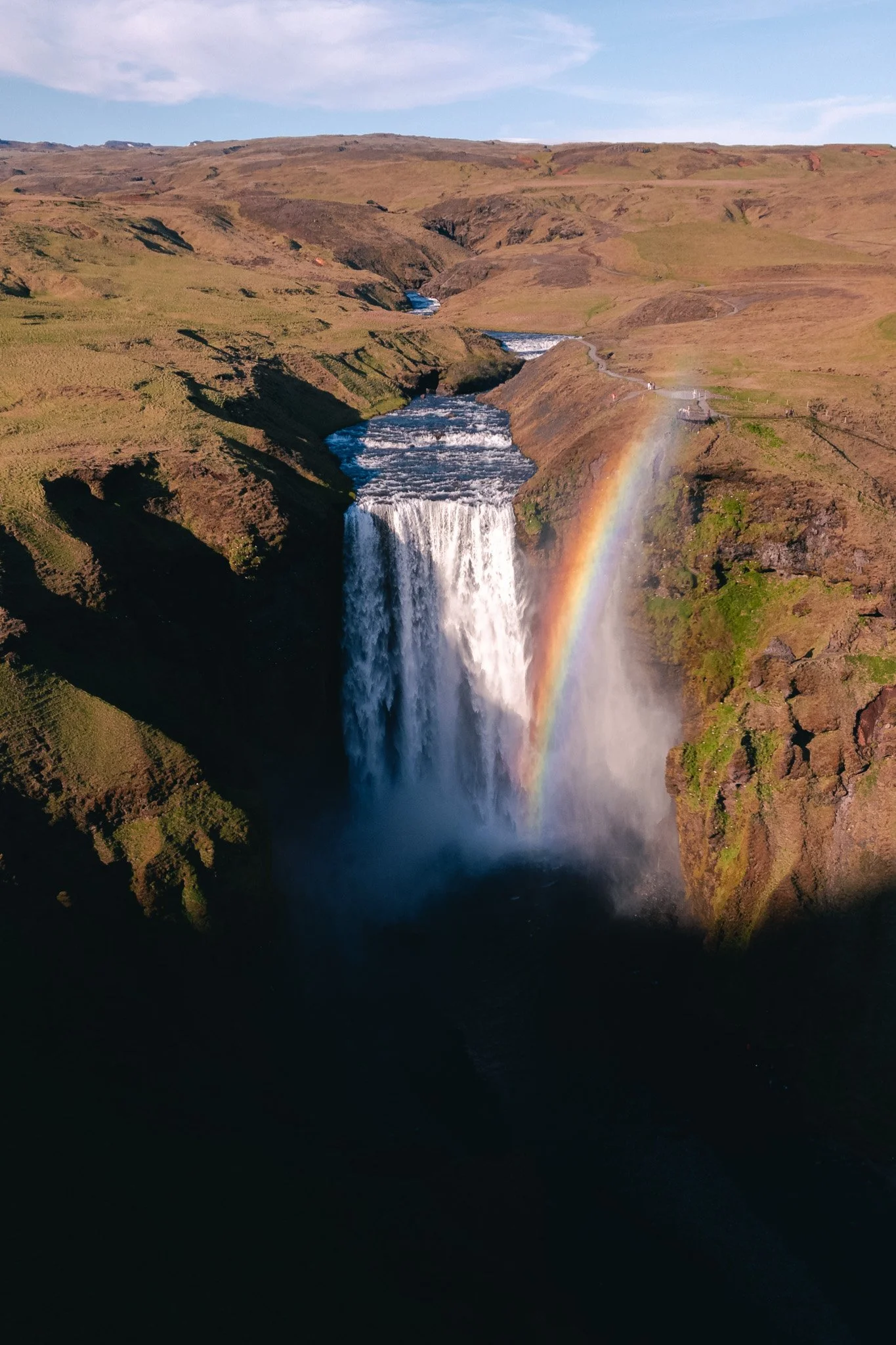 A large waterfall in Iceland with a rainbow forming in the spray, surrounded by green hills and a partly cloudy sky.
