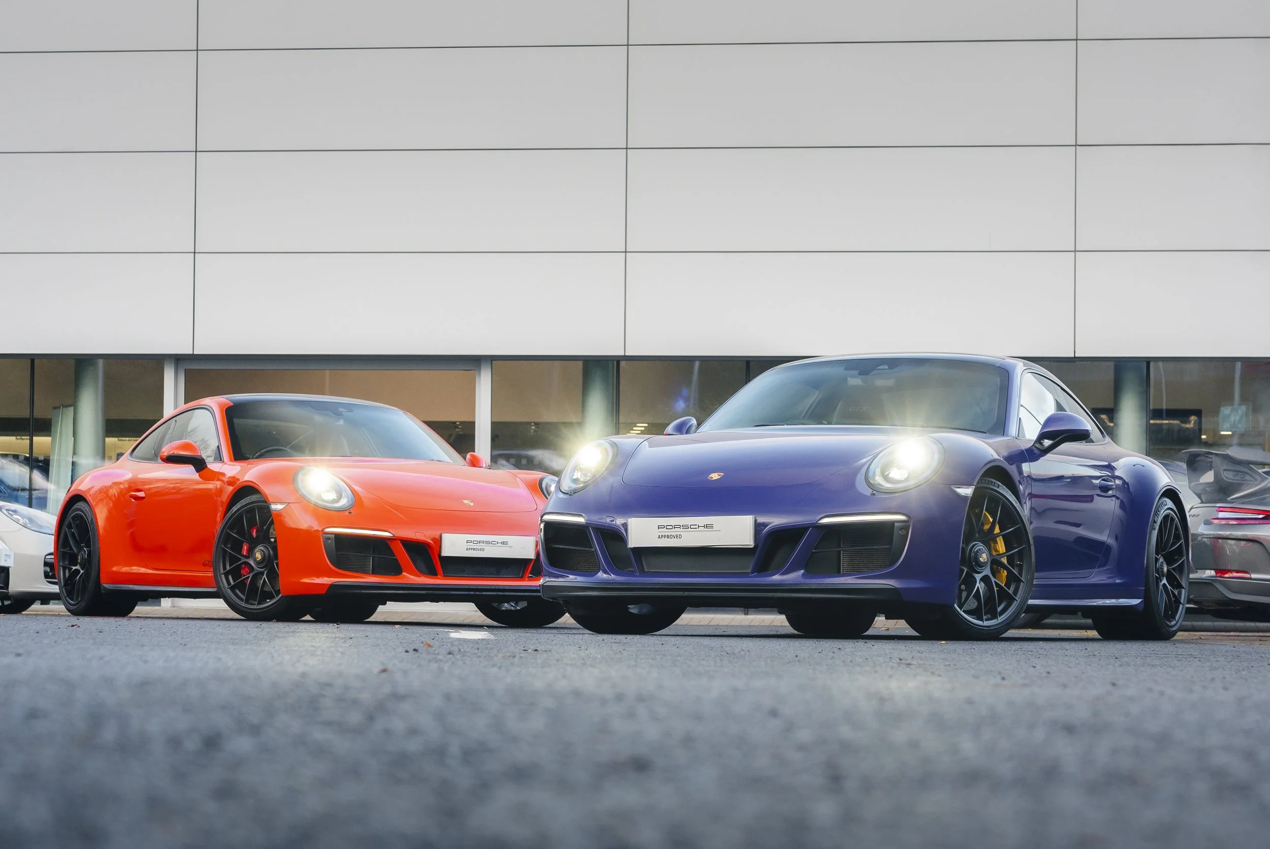 Two Porsche sports cars, one red and one blue, parked outside a dealership. Both cars are sleek with bright headlights on.