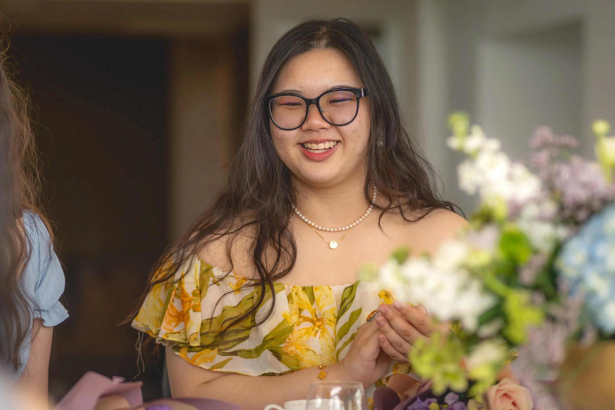 A smiling young woman with glasses, wearing a yellow floral off-shoulder dress and pearl jewelry, stands near a table with flowers, in a bright indoor setting.