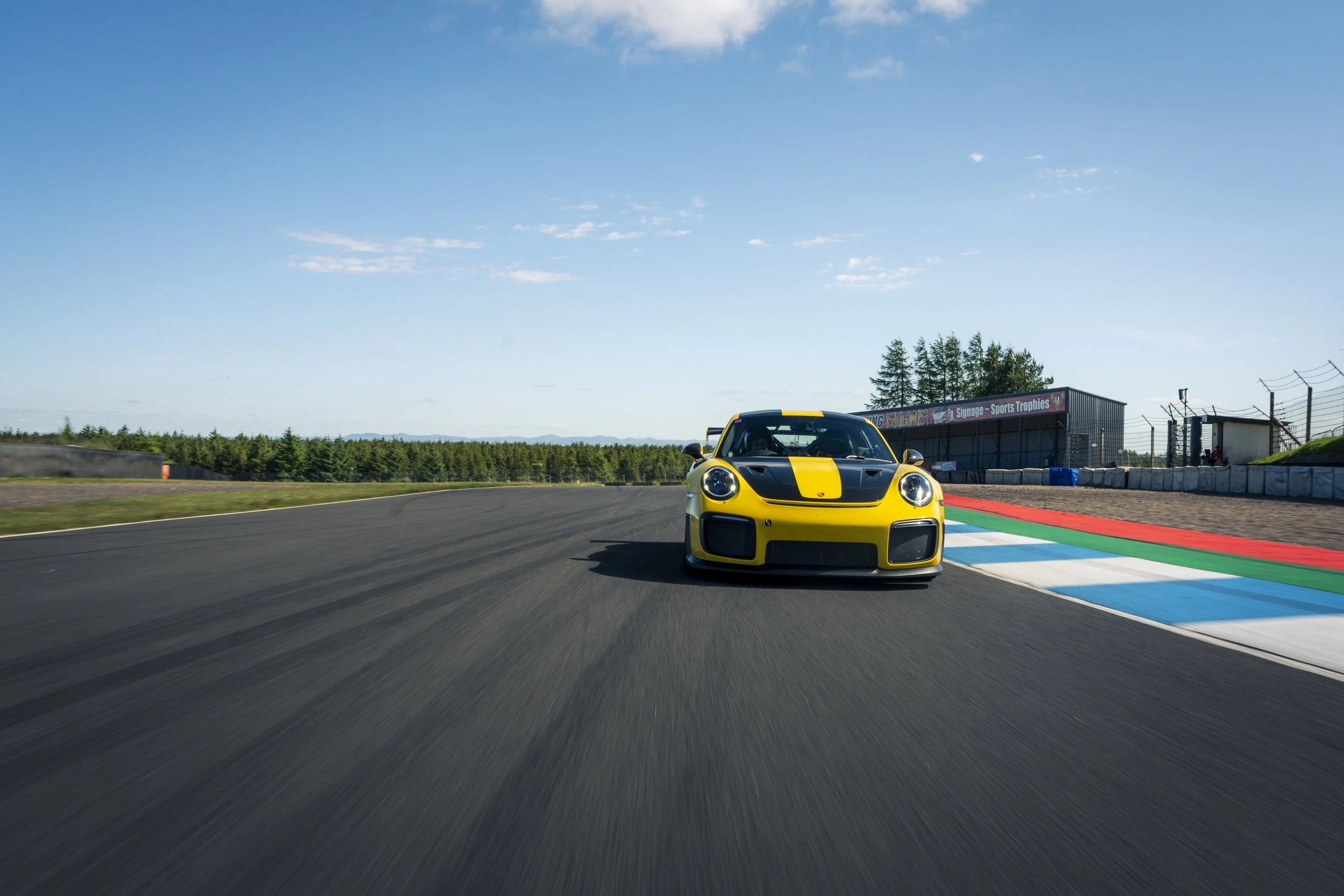 A yellow Porsch GT2RS with black accents racing on a racetrack on a sunny day.