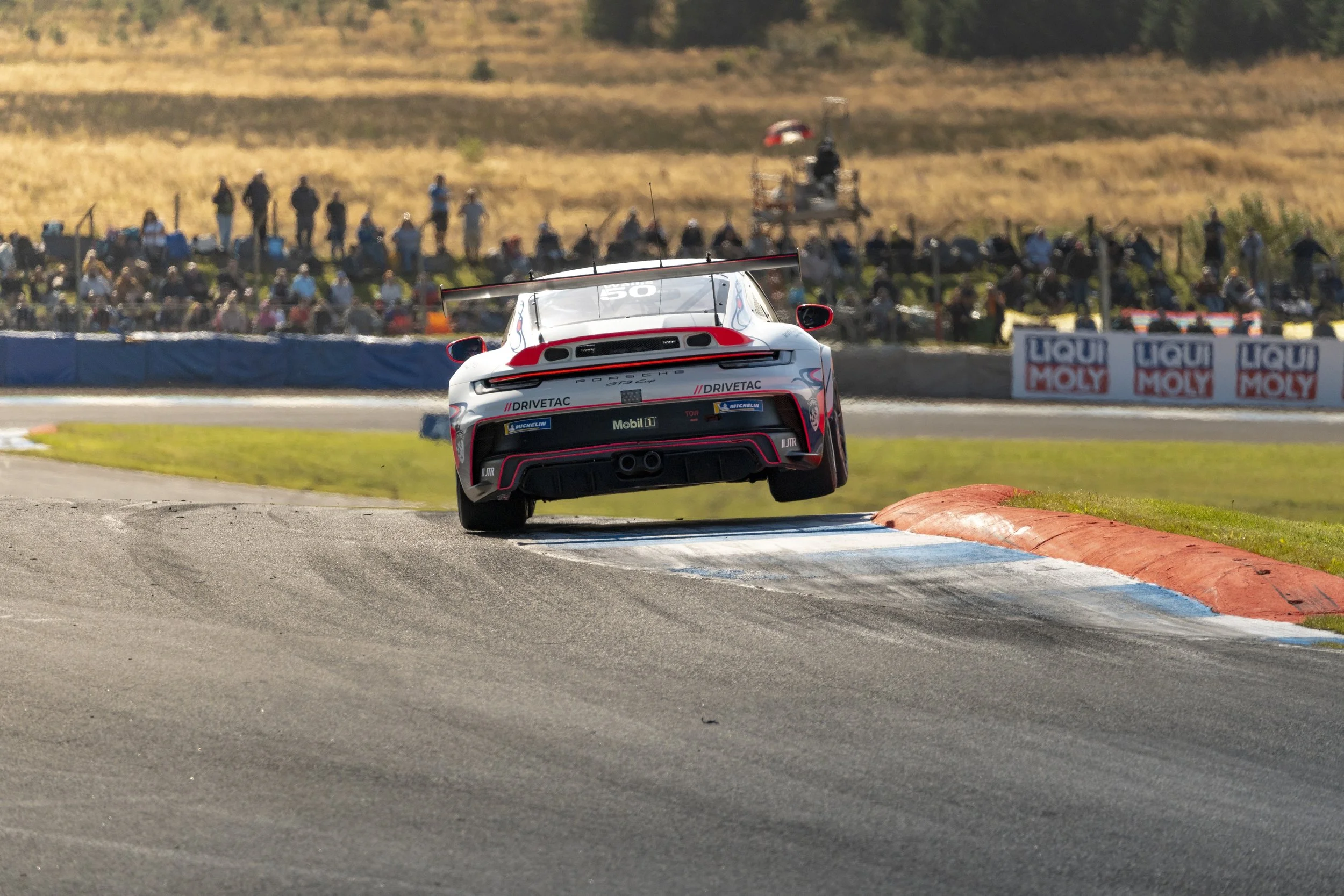 Race car airborne during a race on a track, with spectators watching from the sidelines. Porsche Carrera Cup