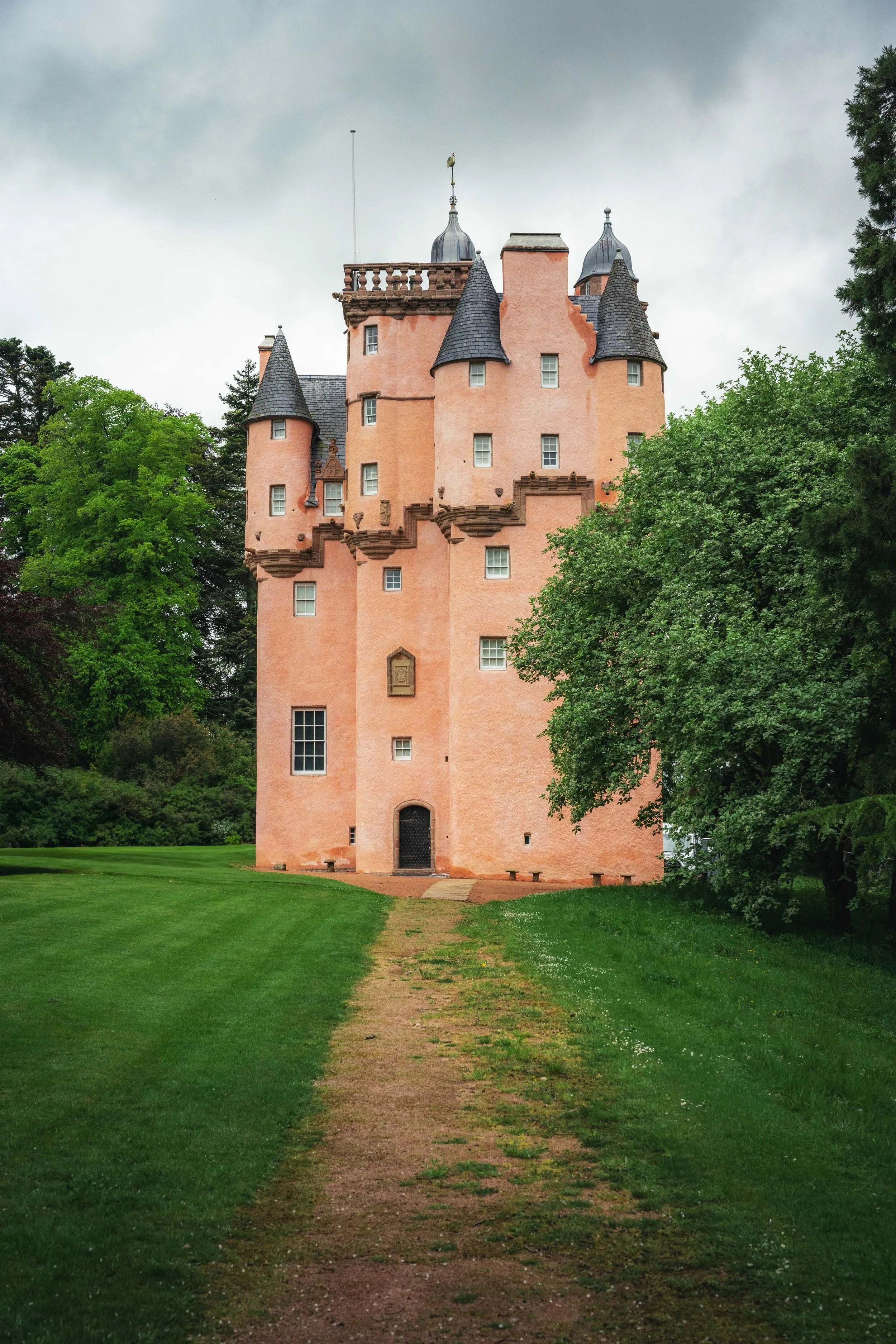 Pink castle with multiple towers on a green lawn, surrounded by trees, under a cloudy sky.