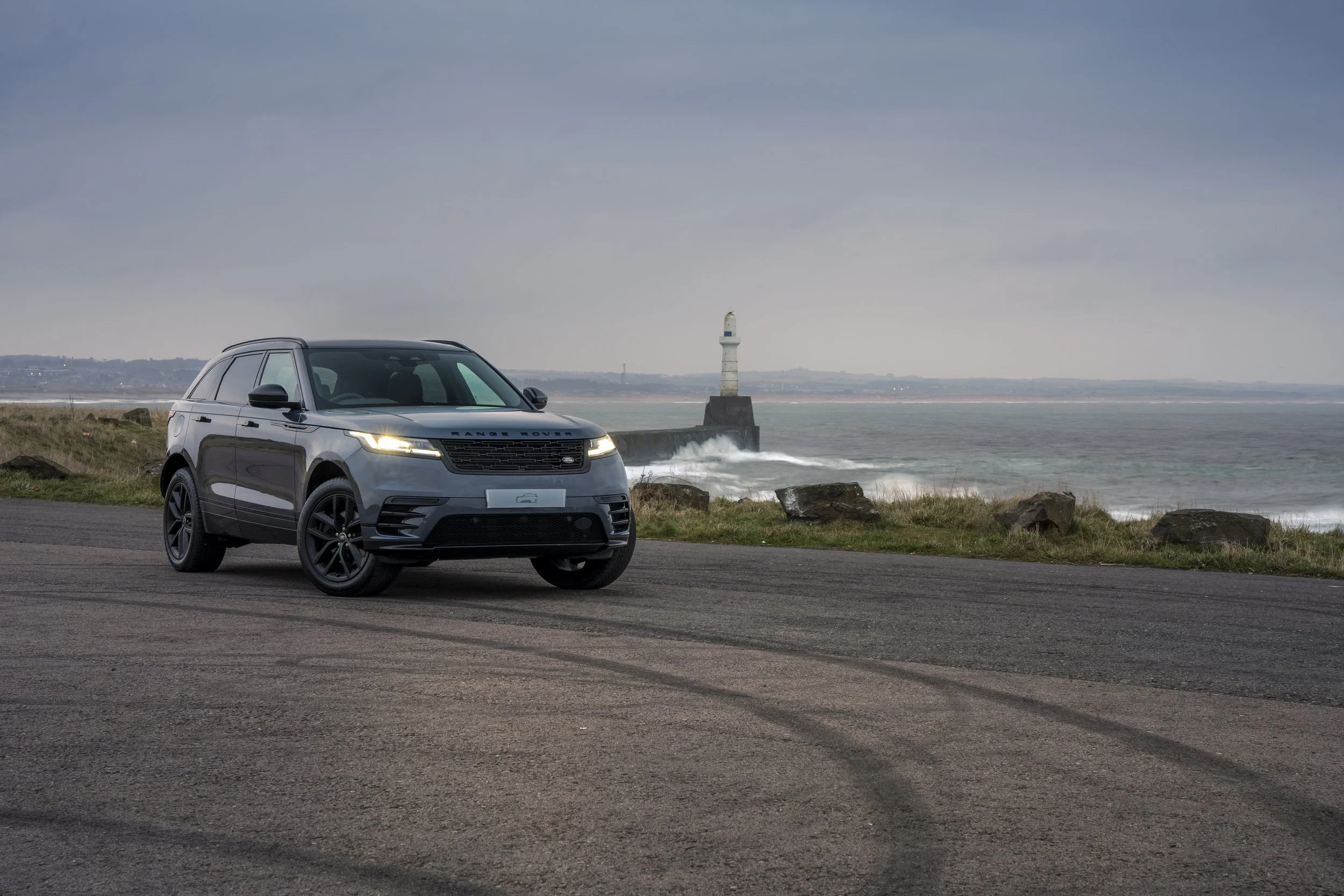 A black Range Rover parked on a coastal road with a lighthouse and rocky shoreline in the background.