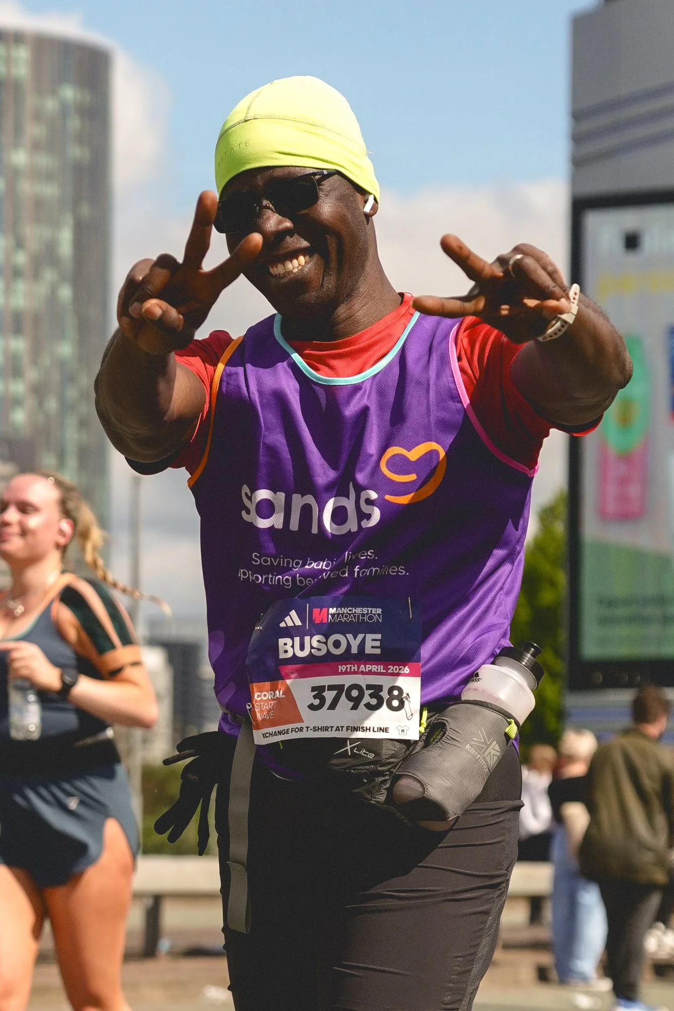 Andre Pena photograph 2026 Manchester Marathon, A smiling man in running gear wearing a purple vest with the word 'sands' on it and a yellow hat, making peace signs with both hands during a marathon race. 