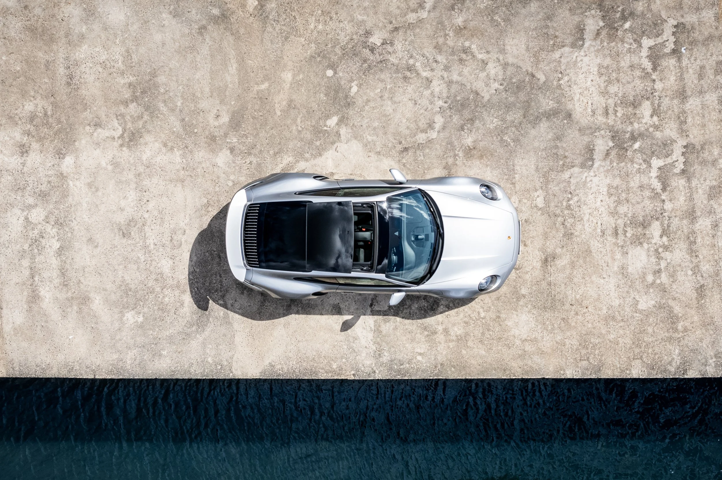 Top-down aerial view of a silver sports car parked on a concrete surface beside a body of water.