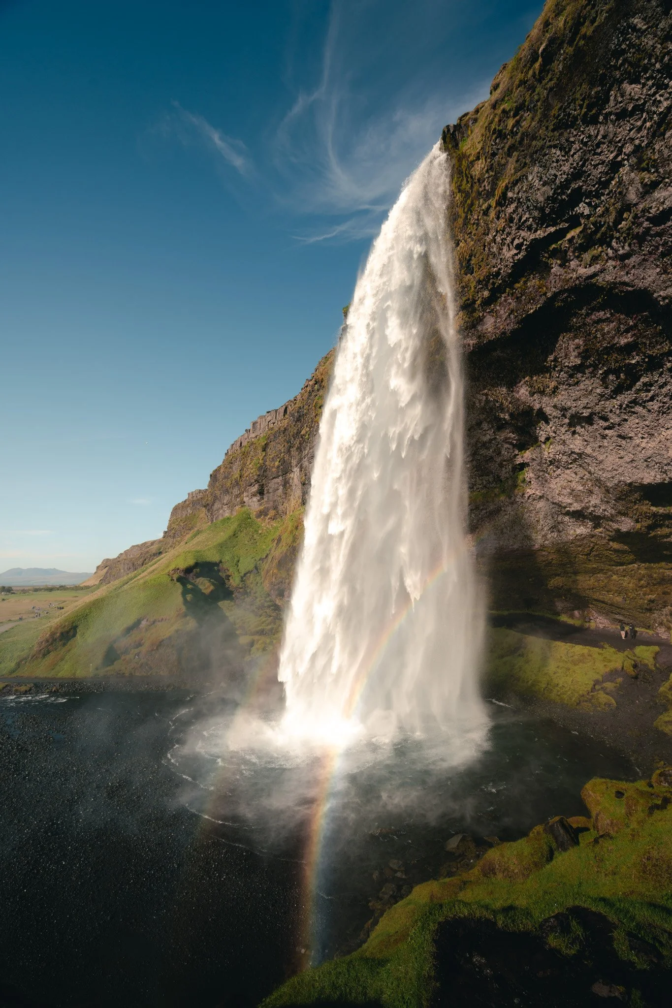 Waterfall cascading over a cliff with a rainbow at the base, green hills, and a blue sky with wispy clouds.
