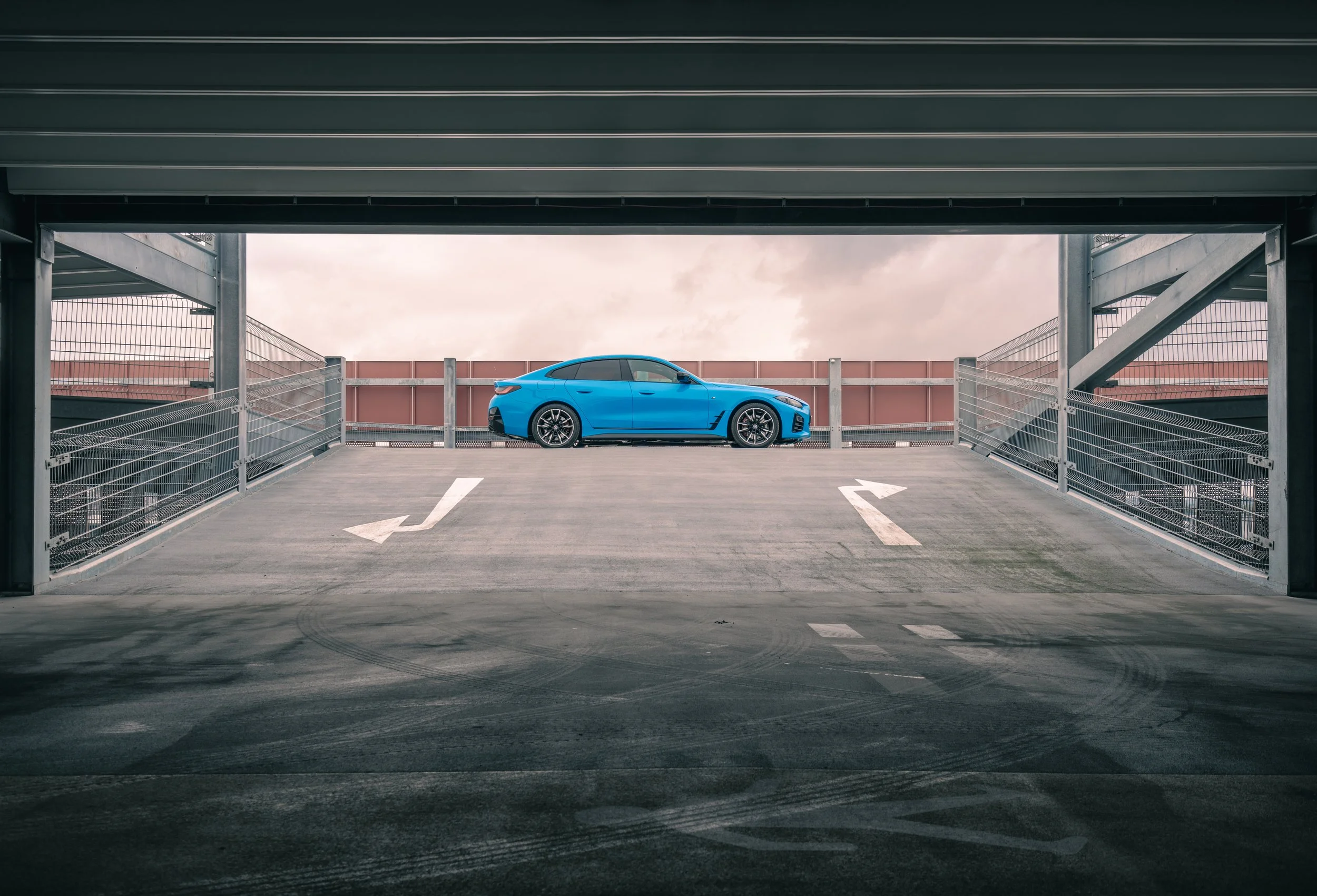 A blue BMW 4 Series parked in an empty multi-level parking garage, viewed from inside the garage with the open ramp leading to the parking space.