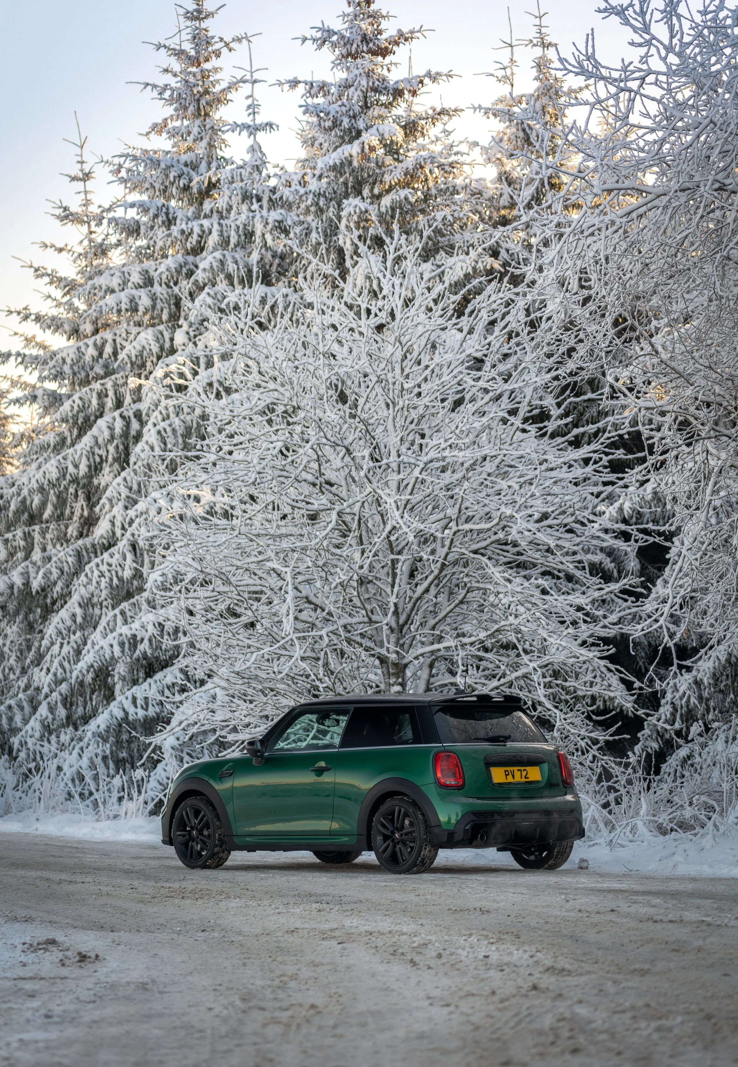 A green compact car parked on a snow-covered road with snow-laden trees in the background during winter.