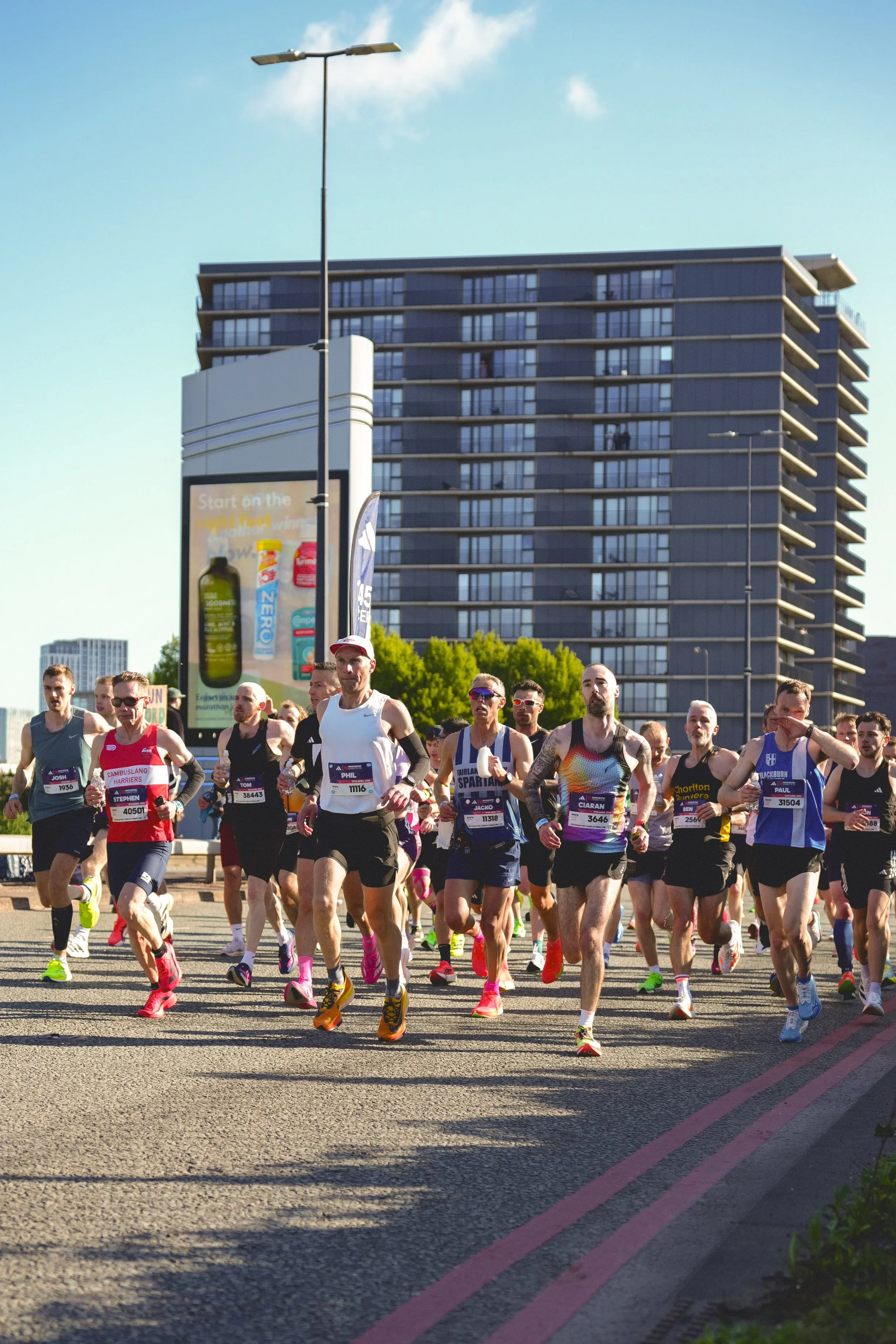 Andre Pena photograph 2026 Manchester Marathon. Group of marathon runners participating in a race on a city street, with a tall building and billboards in the background.