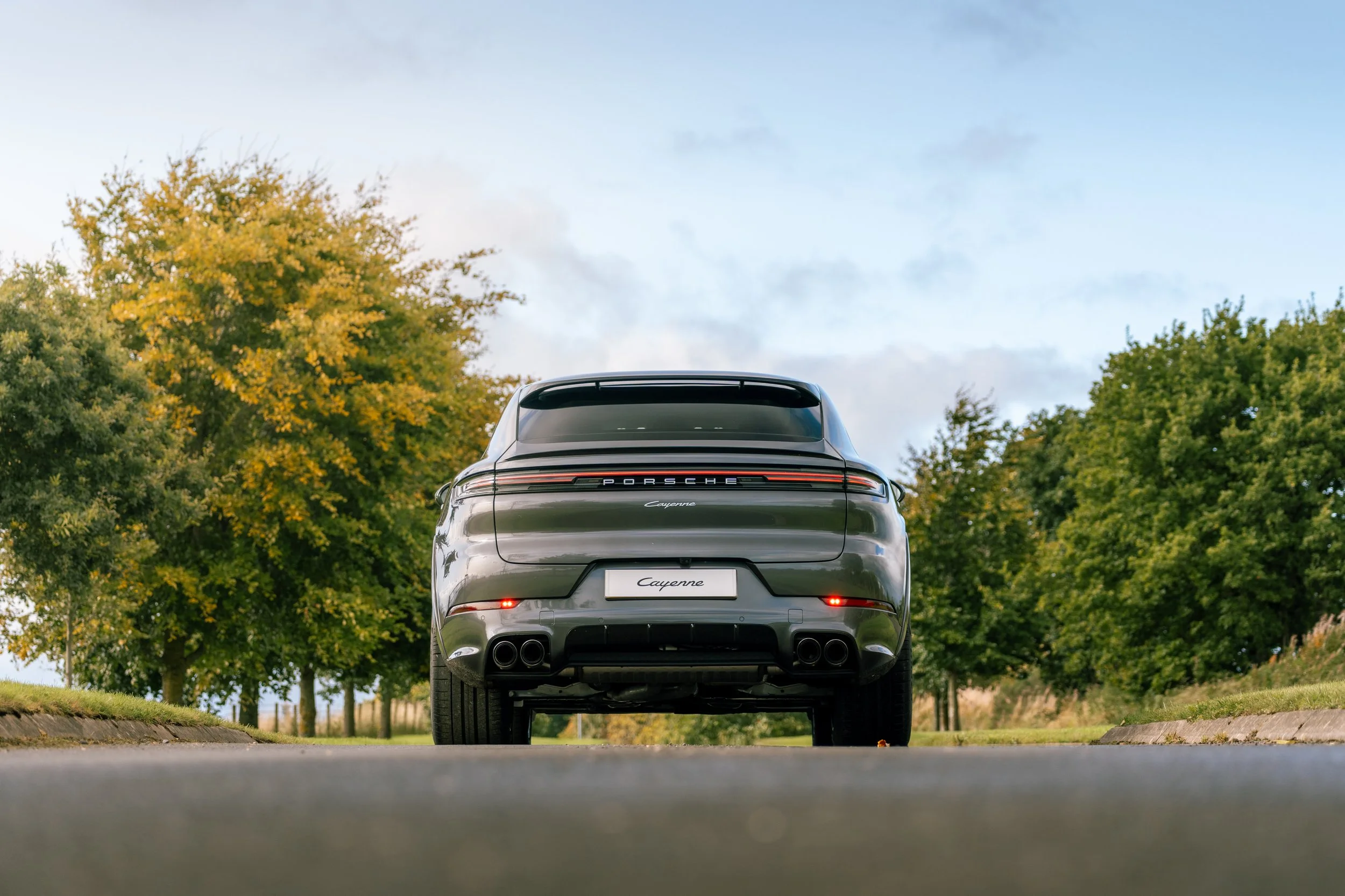 Rear view of a black Porsche Cayenne parked outdoors with green trees and a blue sky in the background.
