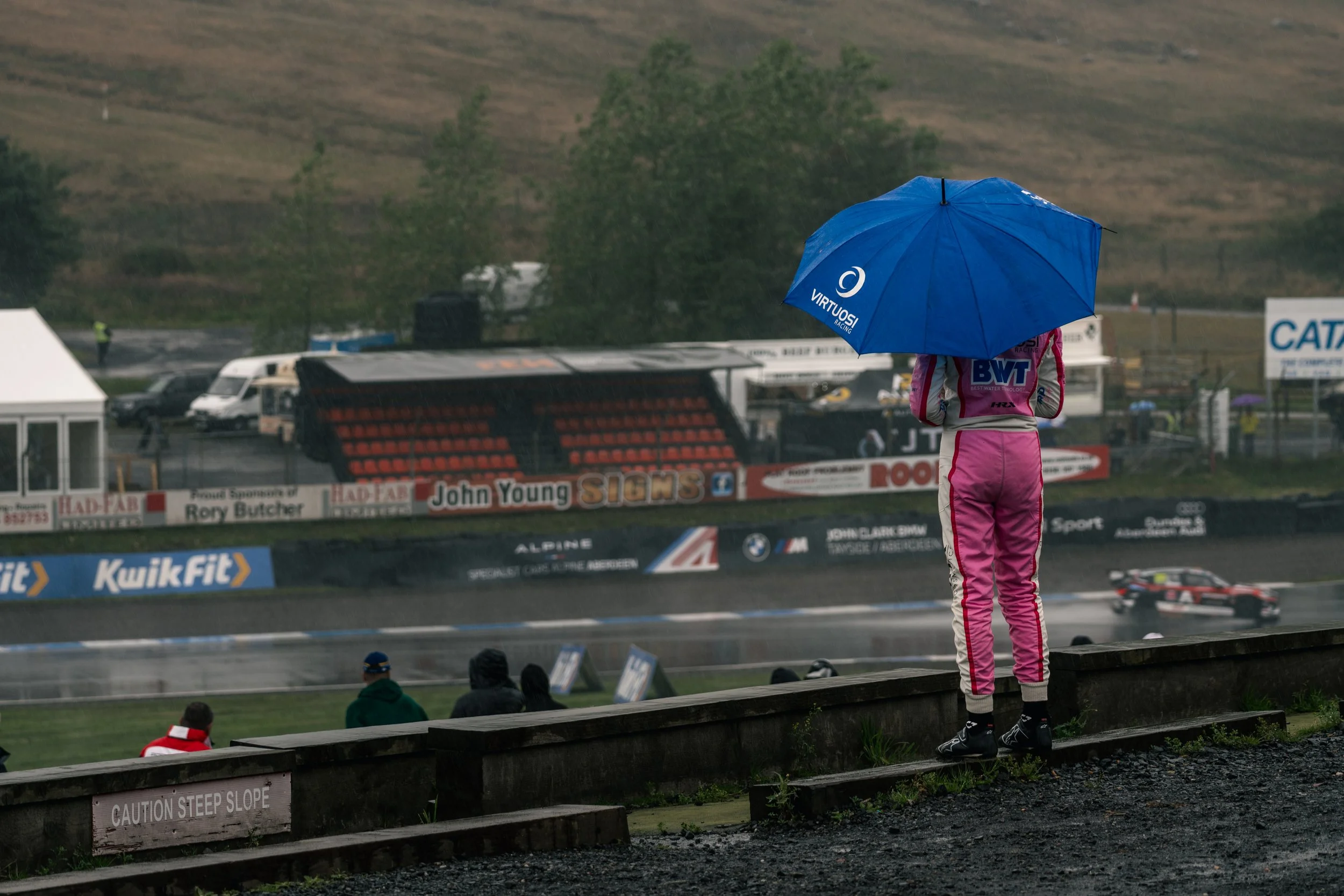 A person wearing a pink racing suit standing on a hillside with a blue umbrella, watching a race on a rainy day at a motorsport track.