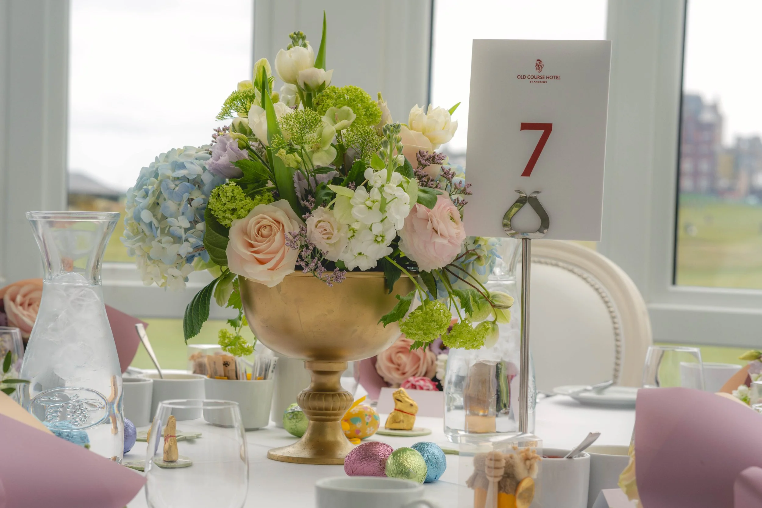 Decorative table centerpiece with a large floral arrangement in a gold vase, featuring white, pink, purple, and green flowers, set at a table with various small decorative items and Easter eggs. A card with the number 7 and the hotel name 'Old Course