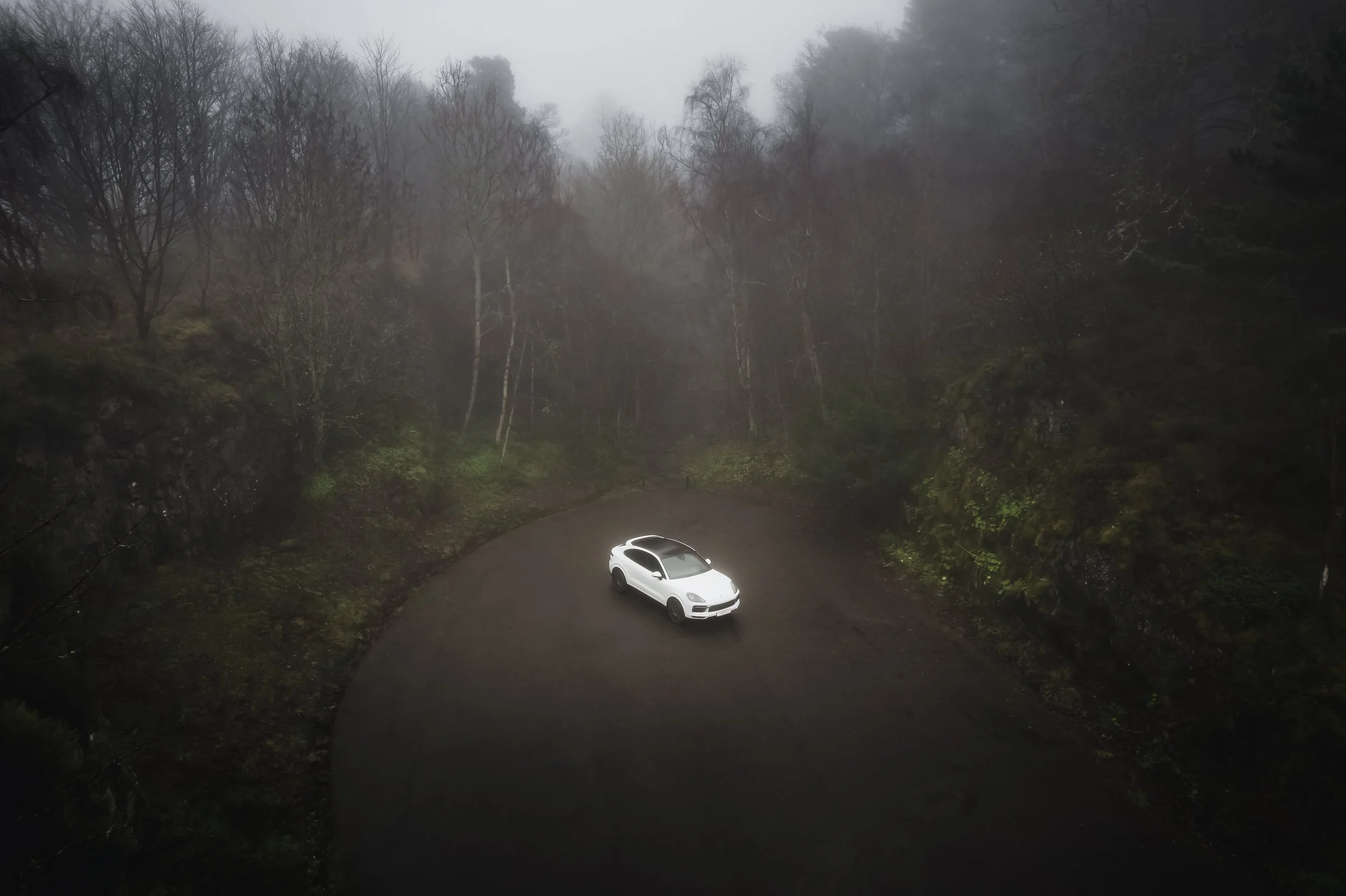 A Porsche Cayenne car parked on a curved, dirt road surrounded by foggy, leafless trees and mossy rocks in a forest.