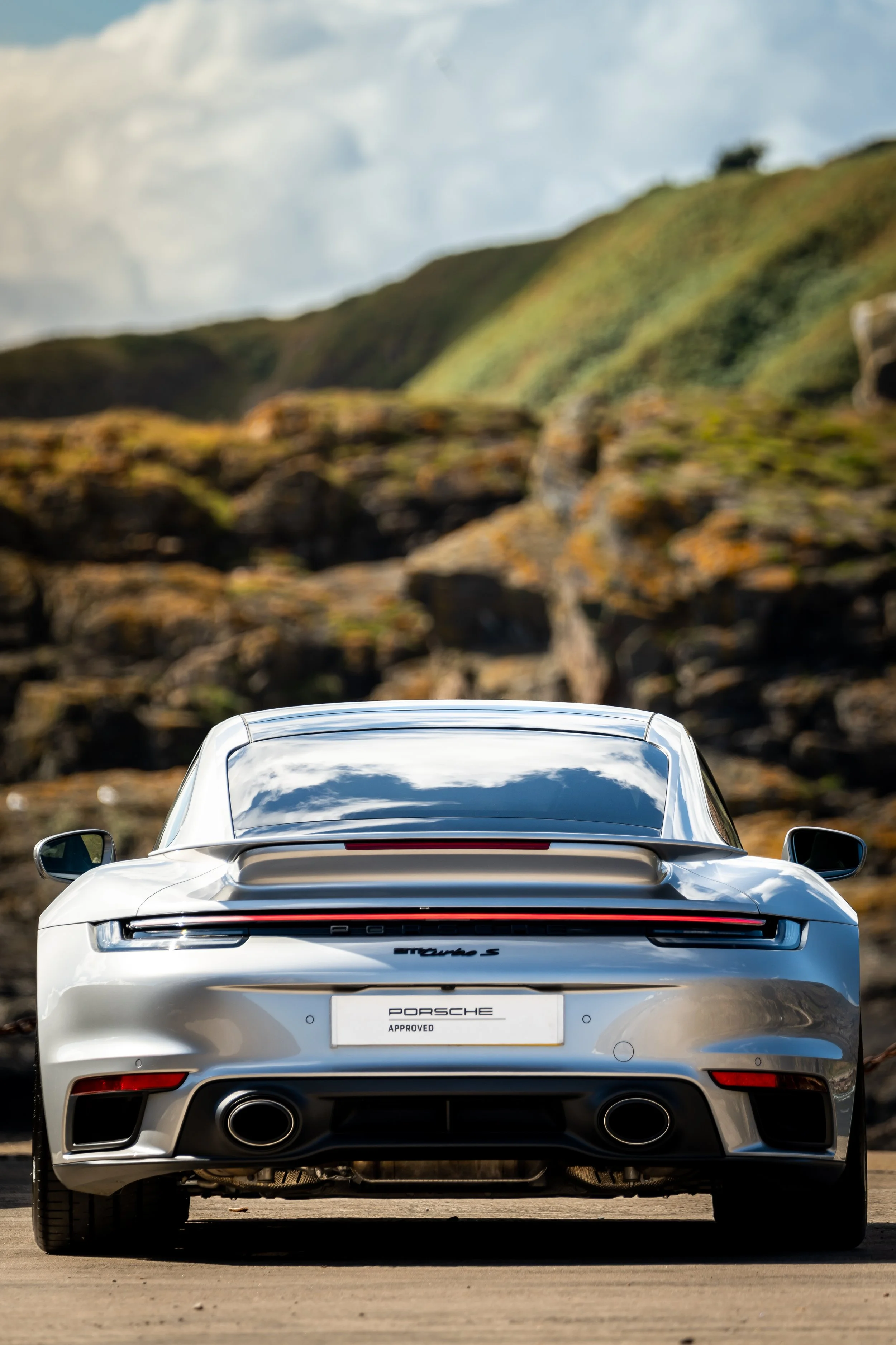 Rear view of a silver Porsche 911 Turbo S sports car parked outdoors with a rocky hillside and green grass in the background.