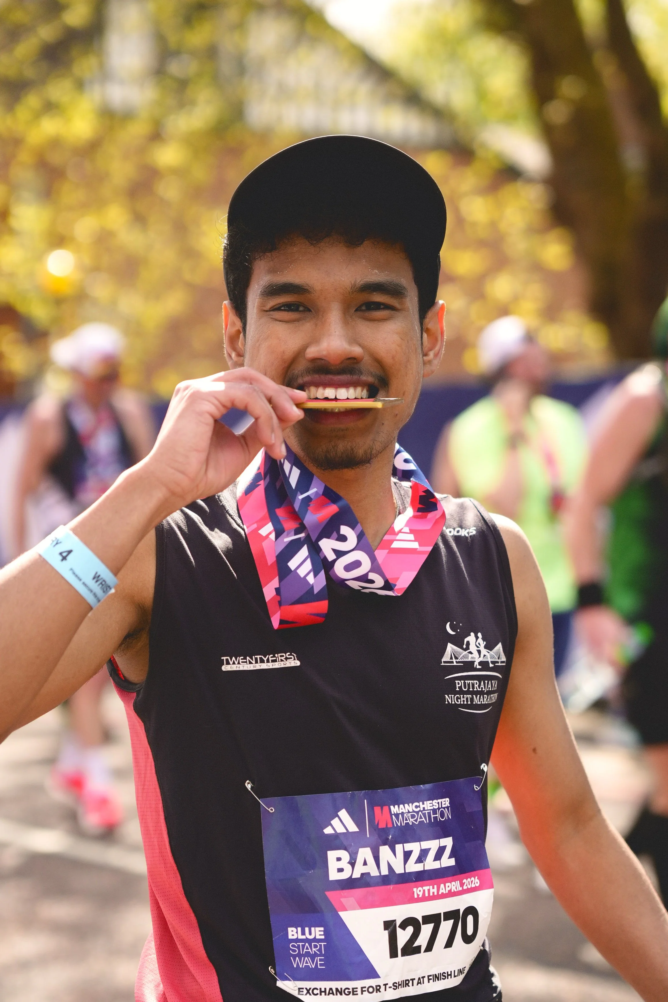 Andre Pena photograph 2026 Manchester Marathon. A man smiling and biting a medal at a marathon event, wearing a black athletic shirt, a medal around his neck. Background shows runners and trees, indicating an outdoor setting.
