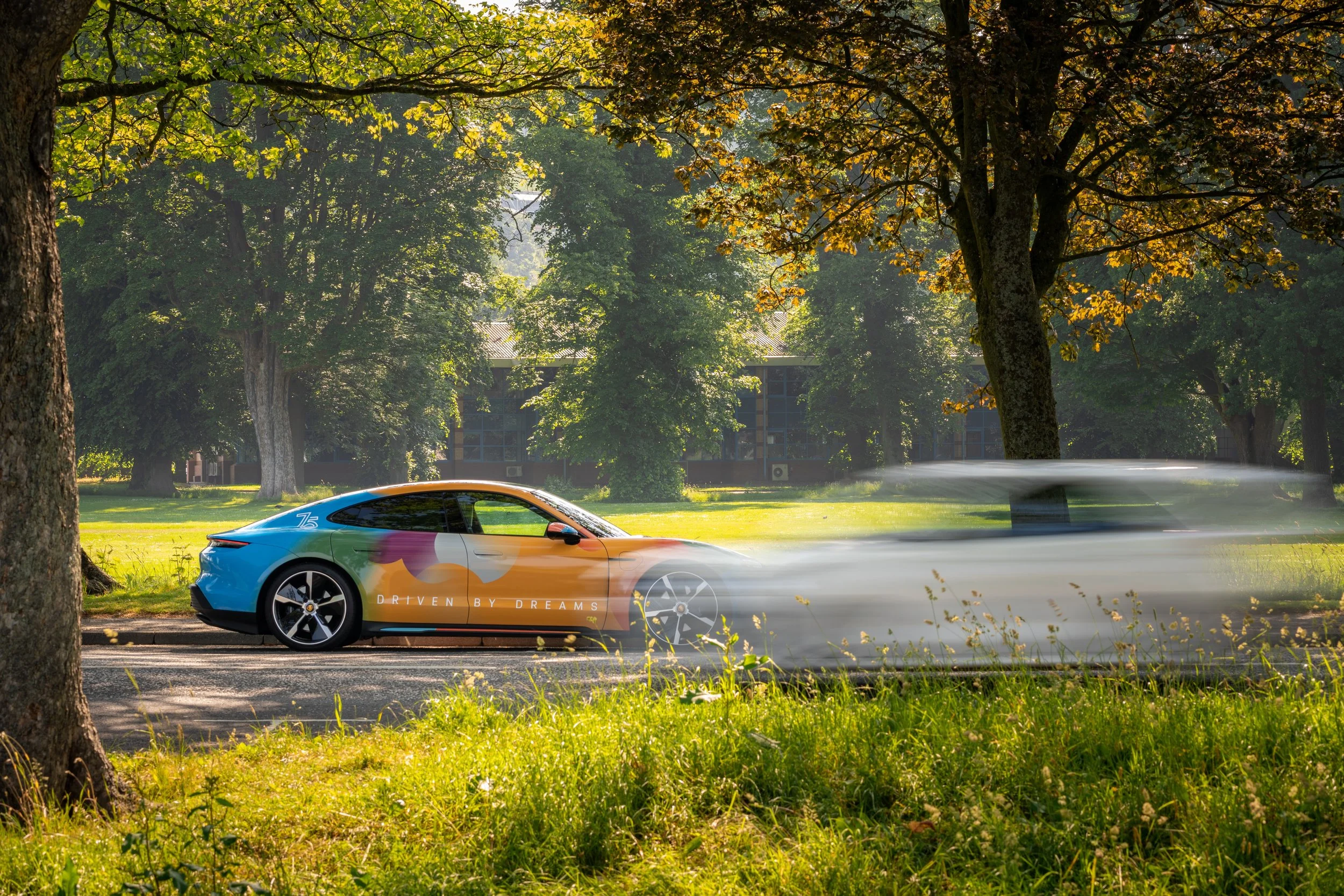 A colorful car with the words "Driven by Dreams" on its side is moving quickly through a park with tall trees and green grass, creating a motion blur effect.