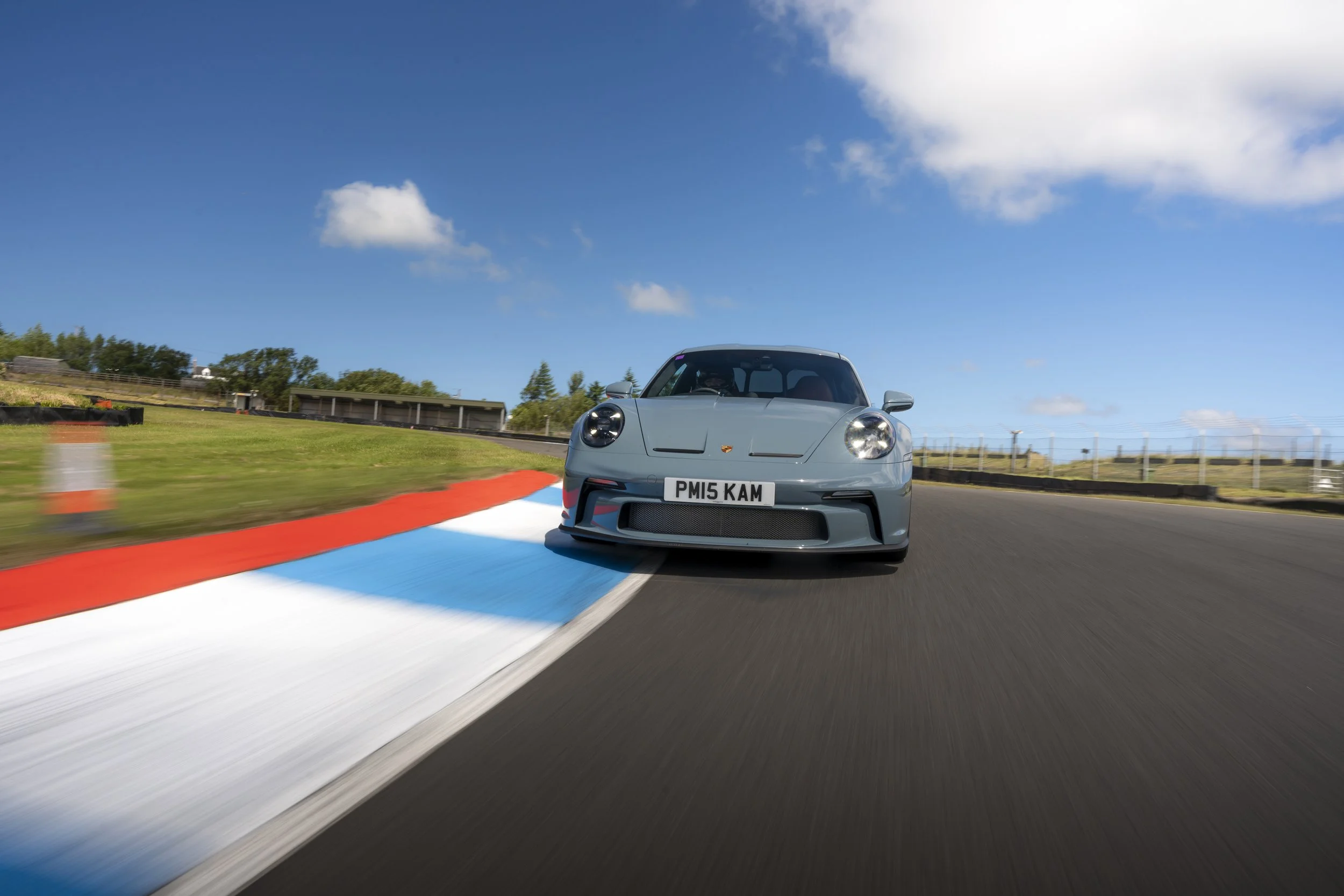 A Porsche 911 ST driving on a race track with striped red, white, and blue curbs under a blue sky with some clouds.